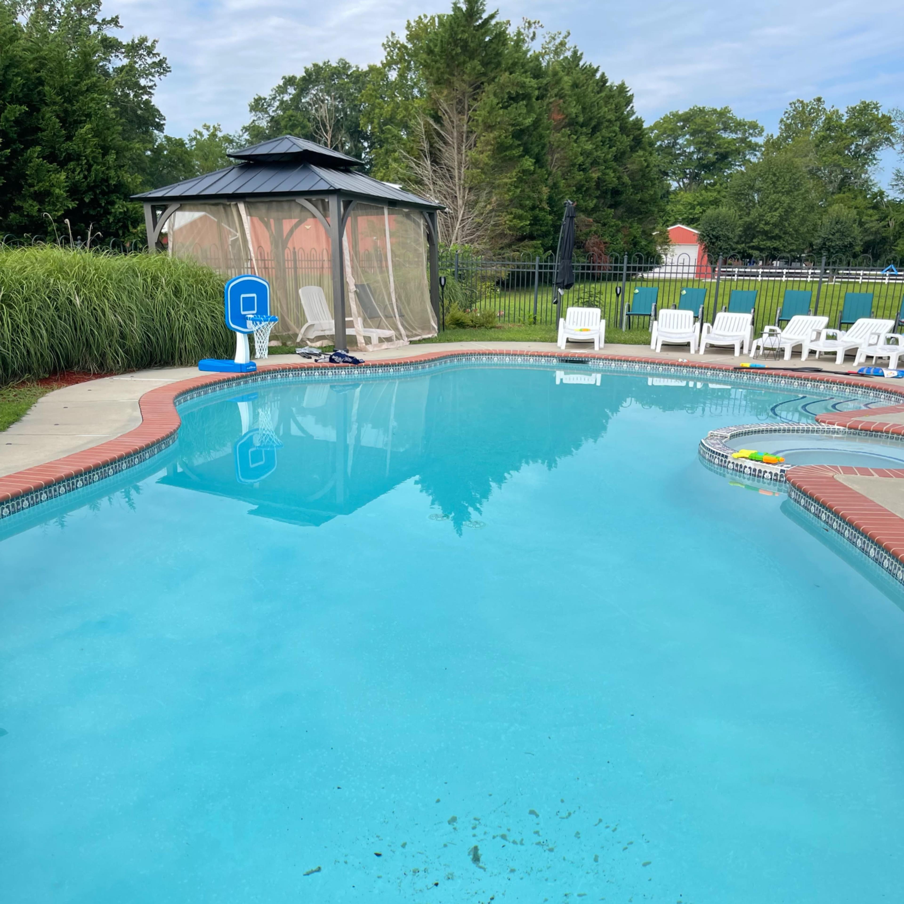 A clear swimming pool is surrounded by chairs and a gazebo in a grassy backyard.