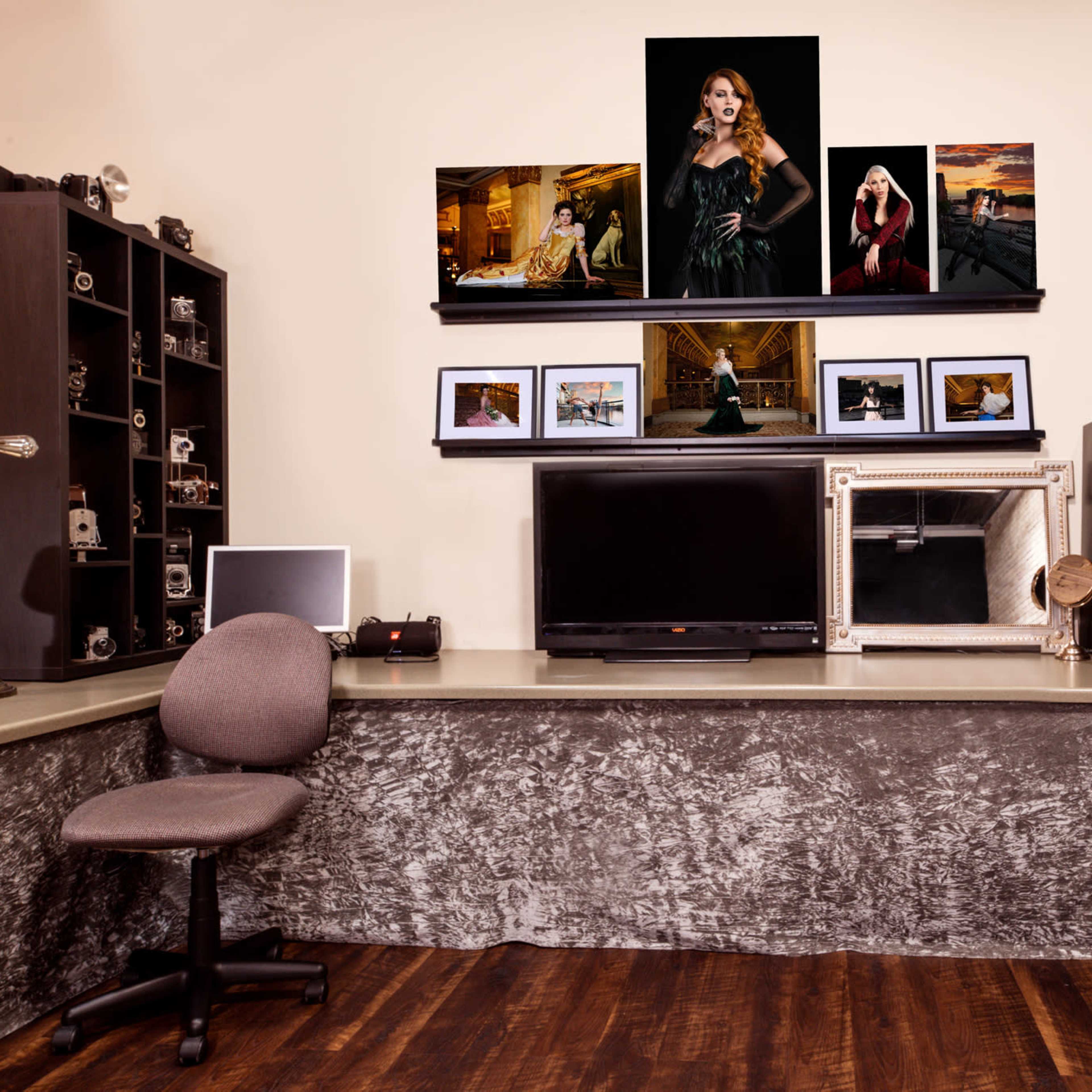 A photography studio with a set of framed photos displayed on the wall above a work surface that includes a television and a small fridge, alongside a chair and various camera equipment.