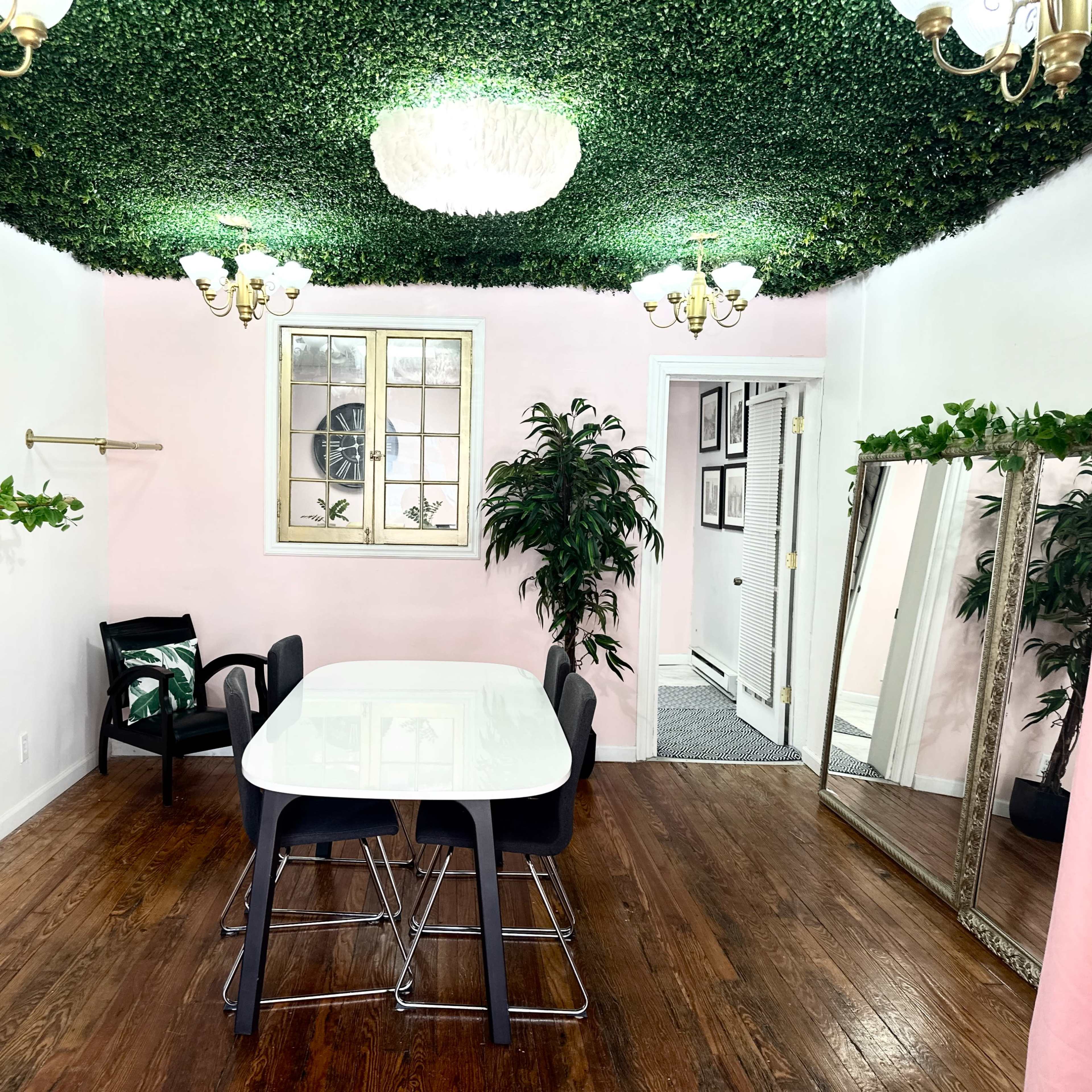 A dining area with a white table and black chairs, surrounded by pink walls, a faux greenery ceiling, and a large mirror.