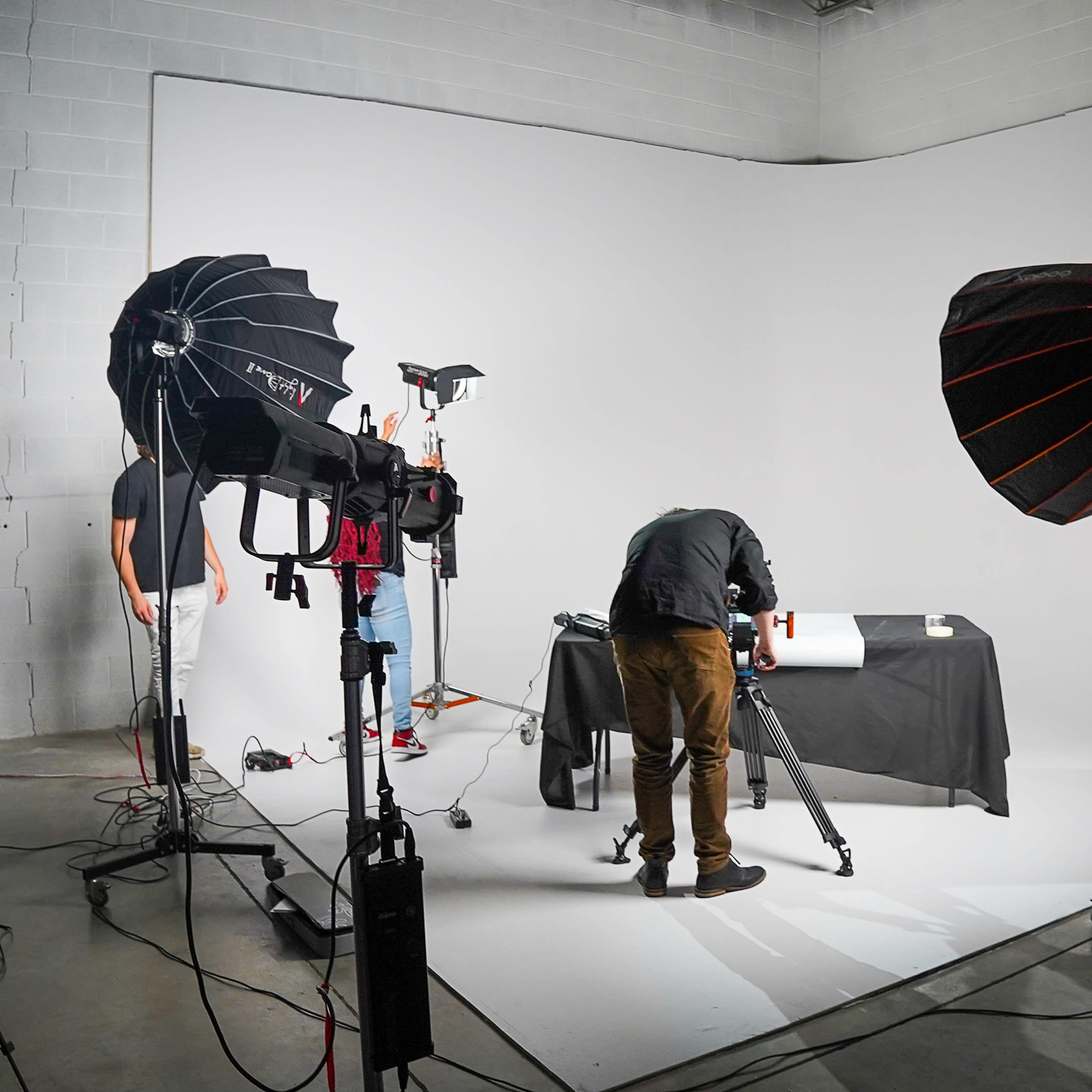 A photography studio setup features a table covered with a black cloth, surrounded by lighting equipment and a person adjusting a camera.