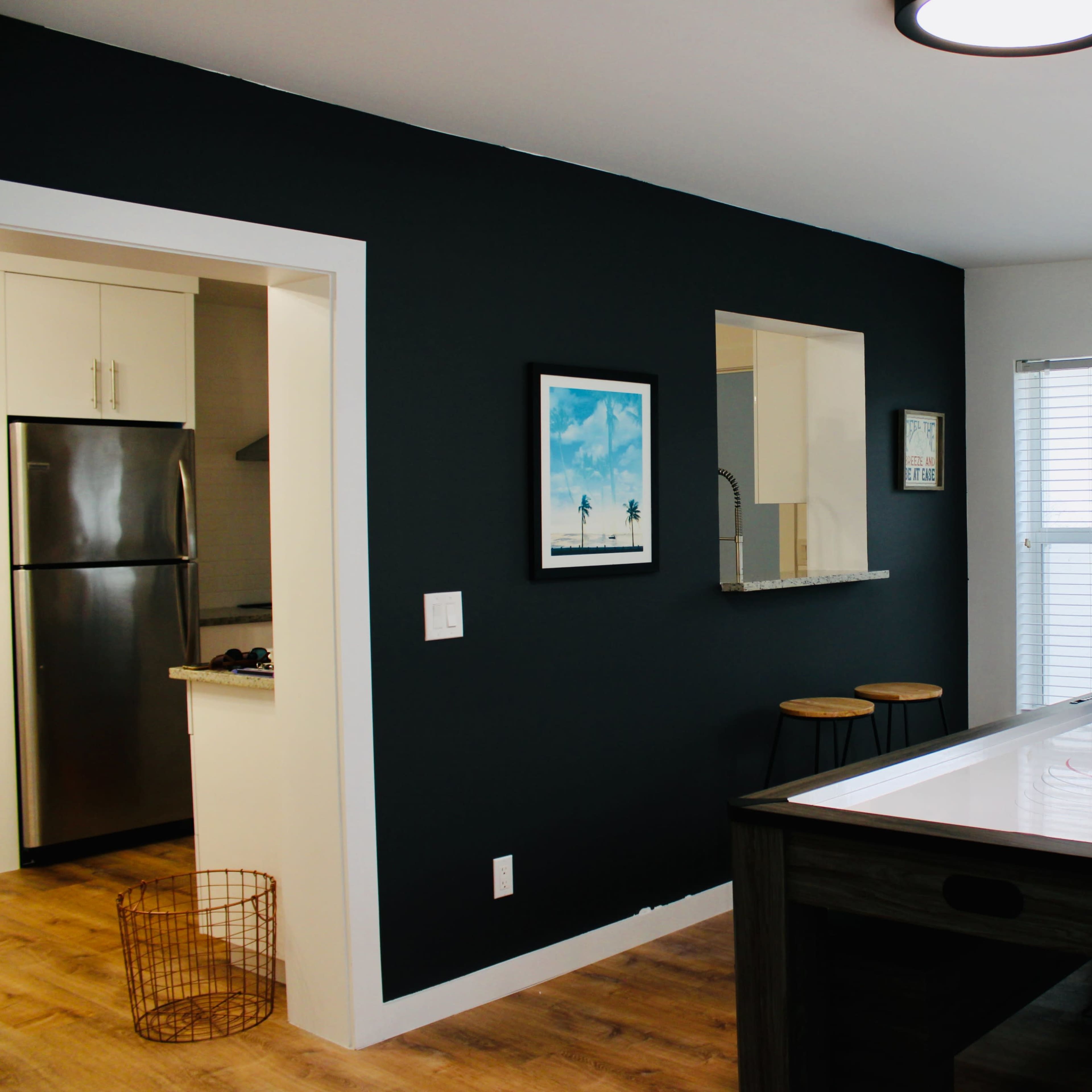 A game room features an air hockey table at the center, with a dark accent wall and an adjoining kitchenette visible through a wide opening.