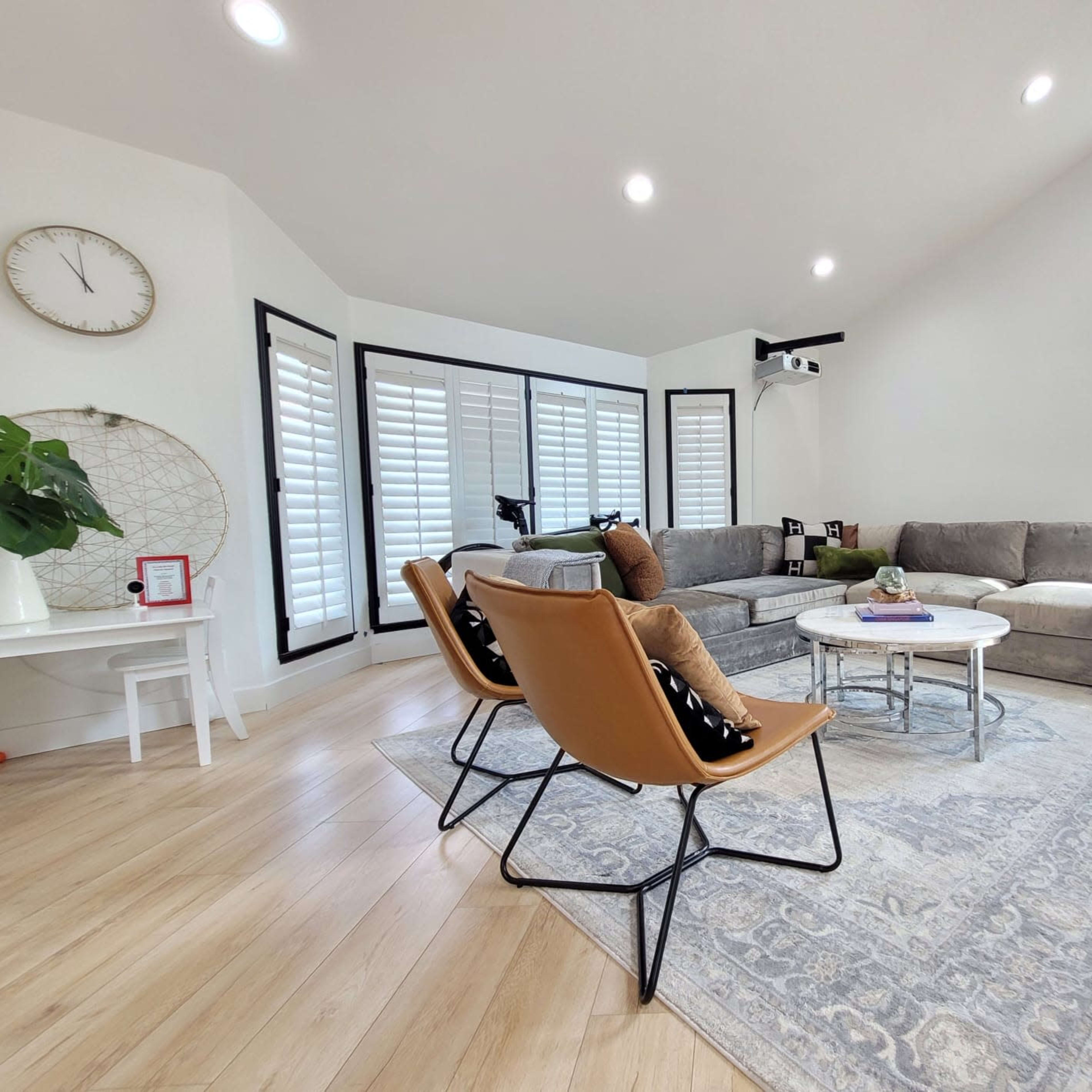 A modern living room with a gray sectional sofa, three leather chairs, a round coffee table, and large windows with plantation shutters.