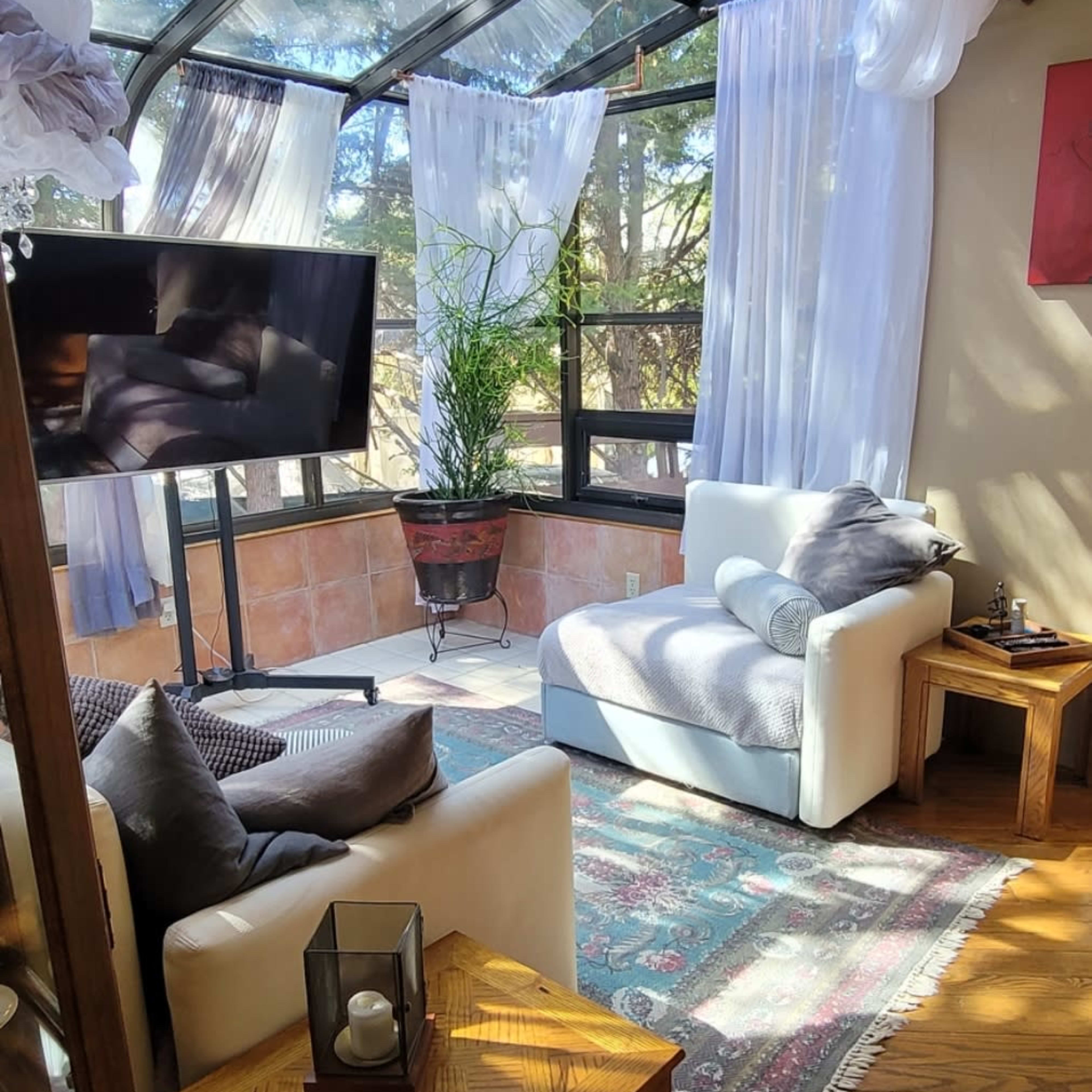 A sunlit sitting area features a glass structure with flowing drapes, a television, a comfortable couch, and a potted plant.