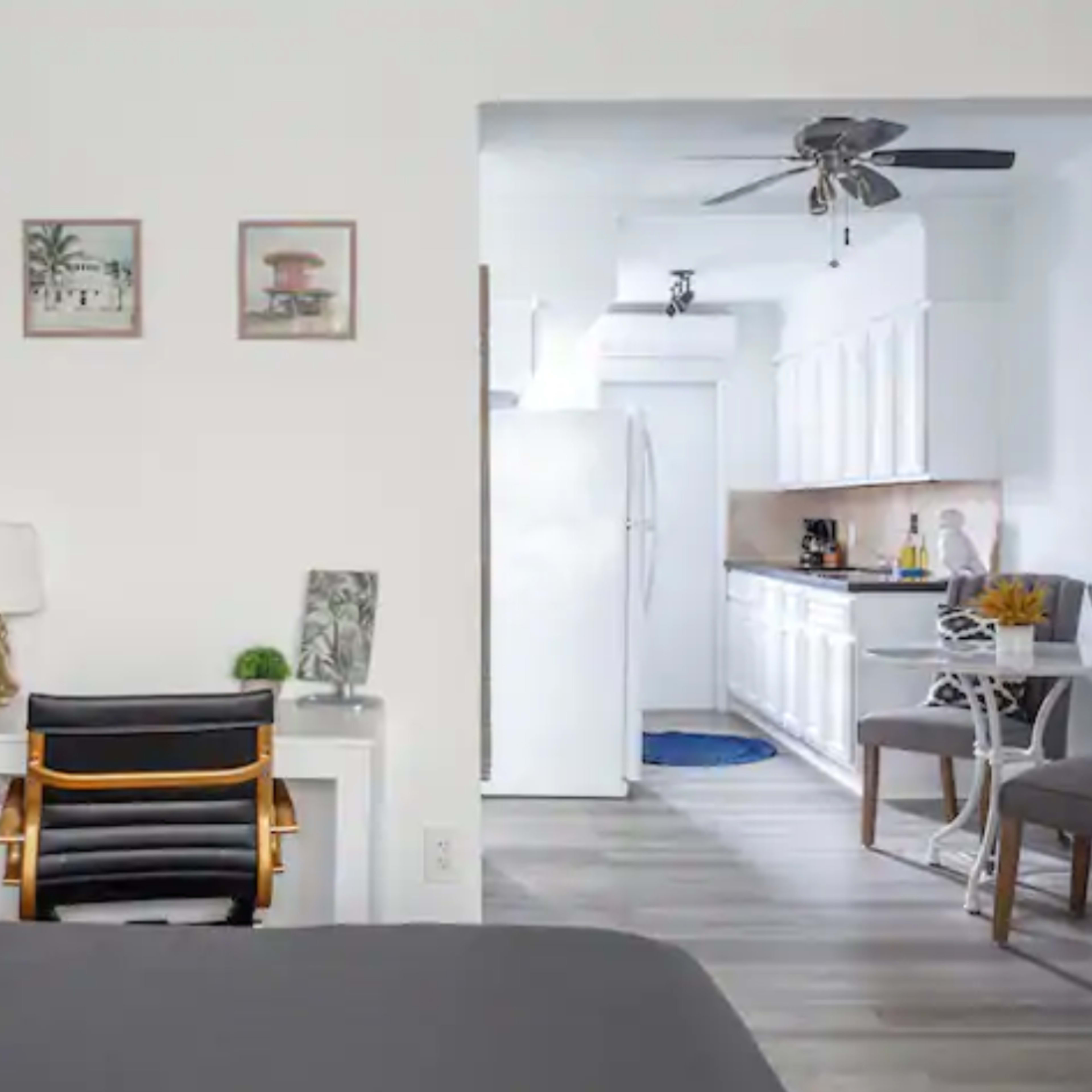 A modern bedroom leading into a kitchen area, featuring a desk and chairs in the foreground, and white cabinetry in the background.