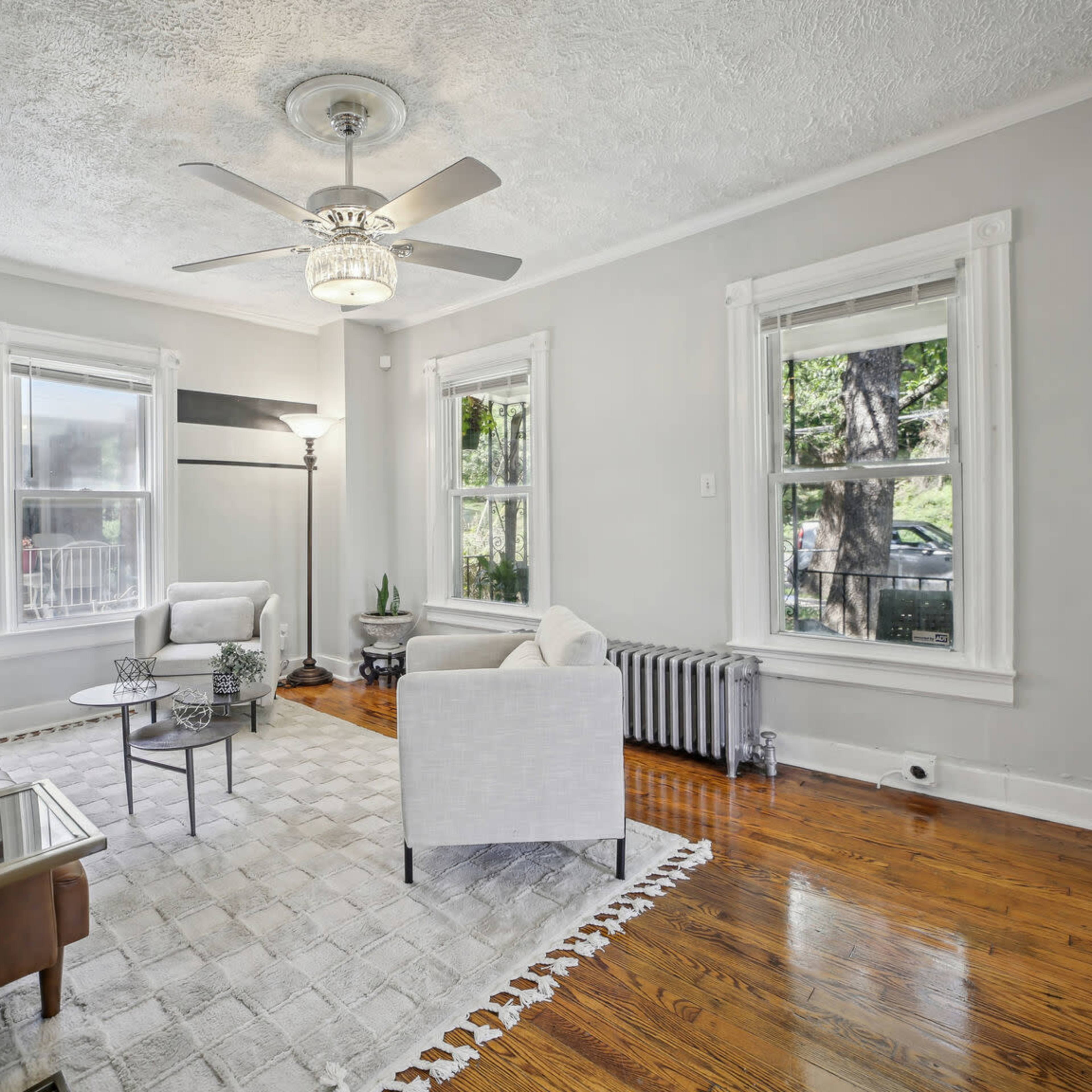 A living room features two armchairs and a leather sofa, with large windows and a ceiling fan above.