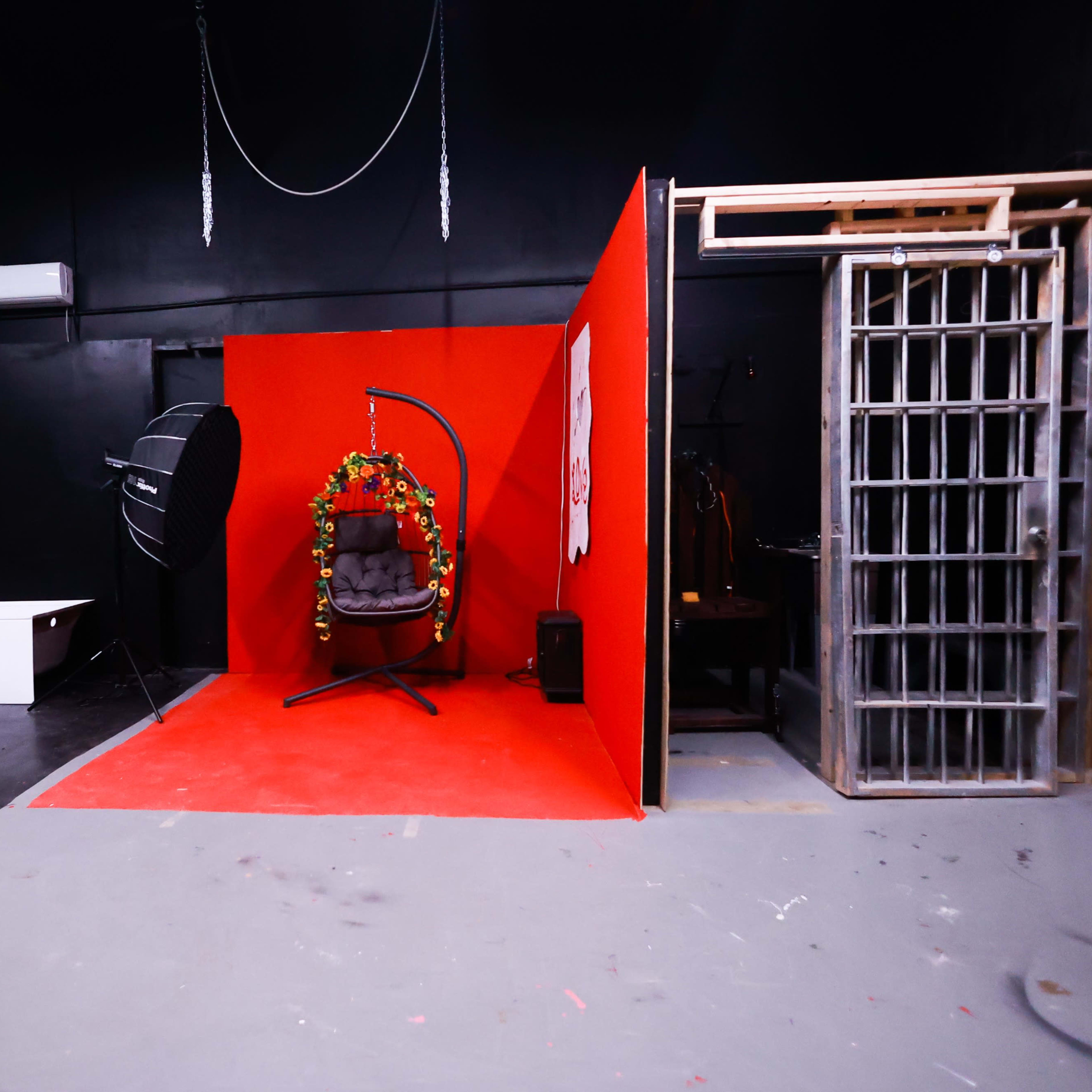 A studio space featuring a red backdrop with a hanging chair decorated with foliage, a lighting setup, and a metal cage-like structure.