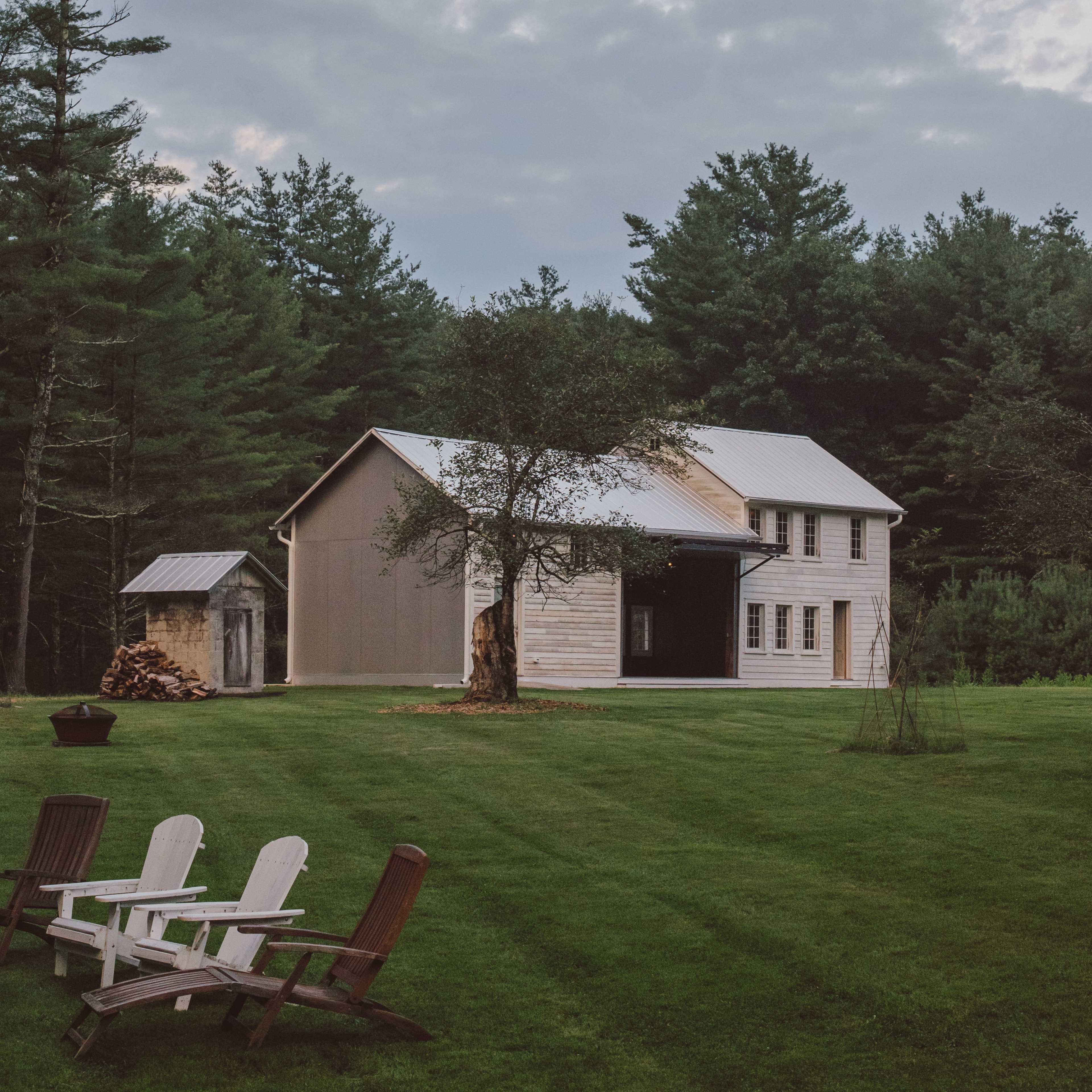 A two-story house with a metal roof and wooden exterior is situated on a grassy area surrounded by trees, with a small shed and stacked firewood nearby.