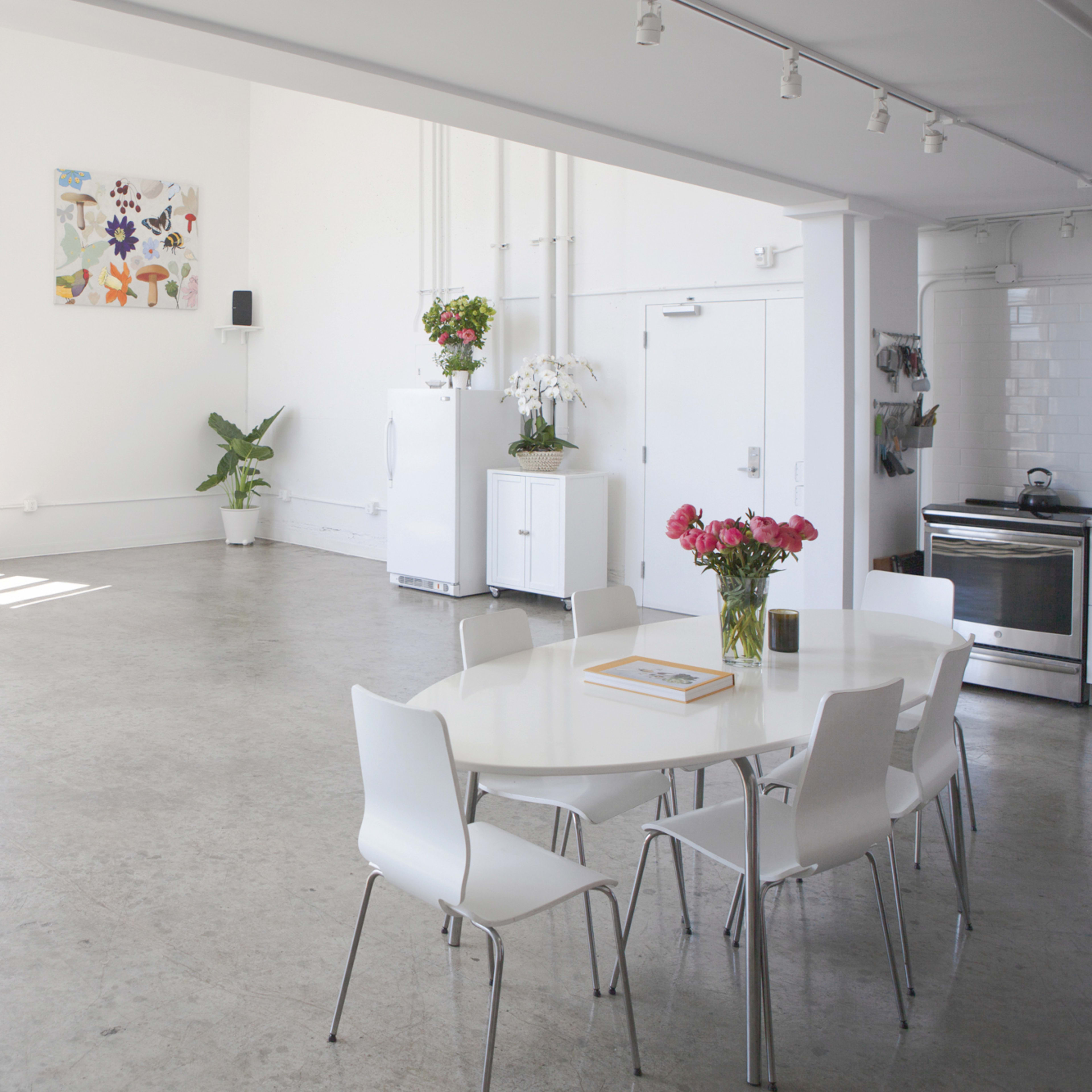 A bright, spacious interior featuring a circular dining table with white chairs, a kitchen area, and large windows allowing natural light to fill the room.