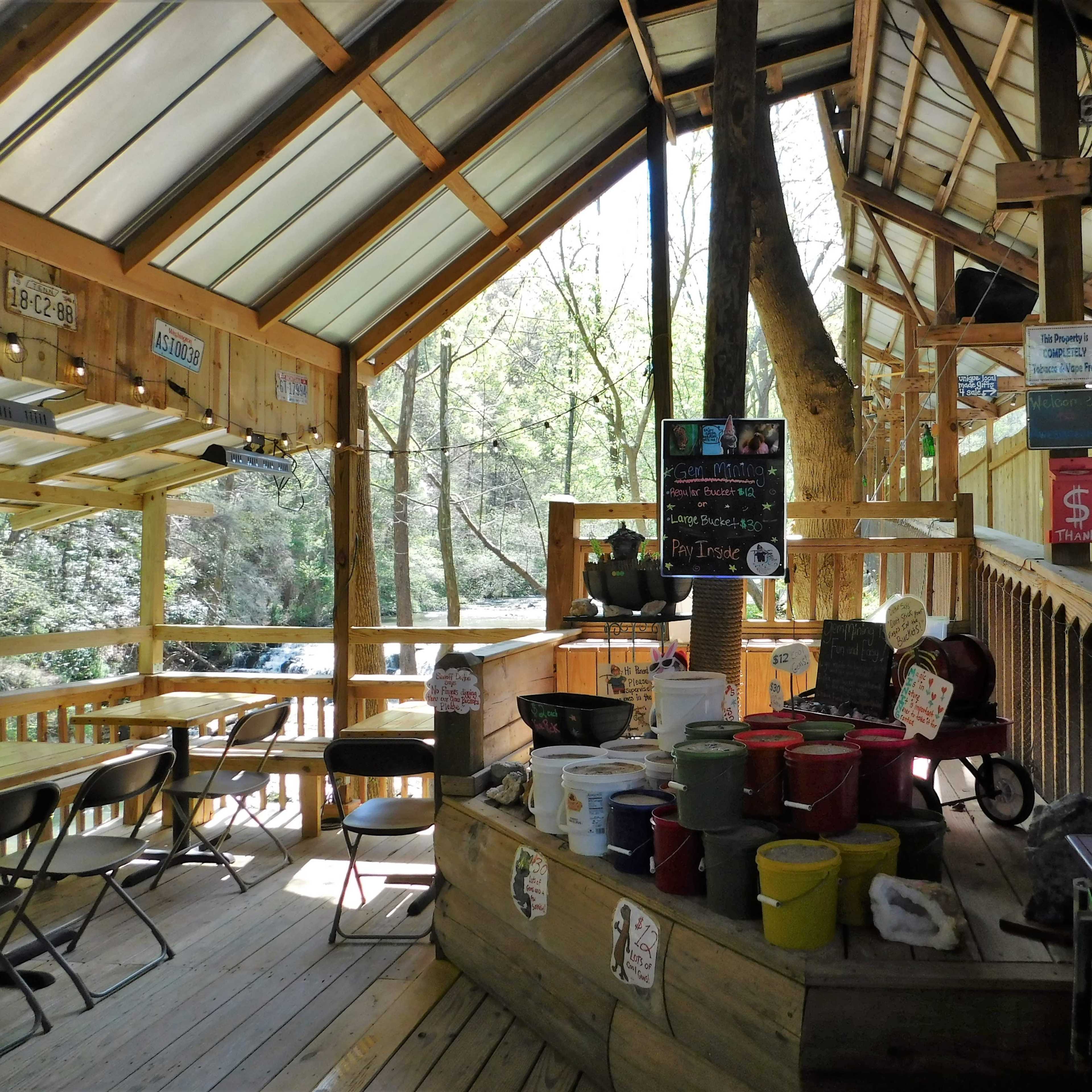 A wooden interior of a rustic café with a counter displaying colorful pots and signs, surrounded by tall trees visible through large windows.