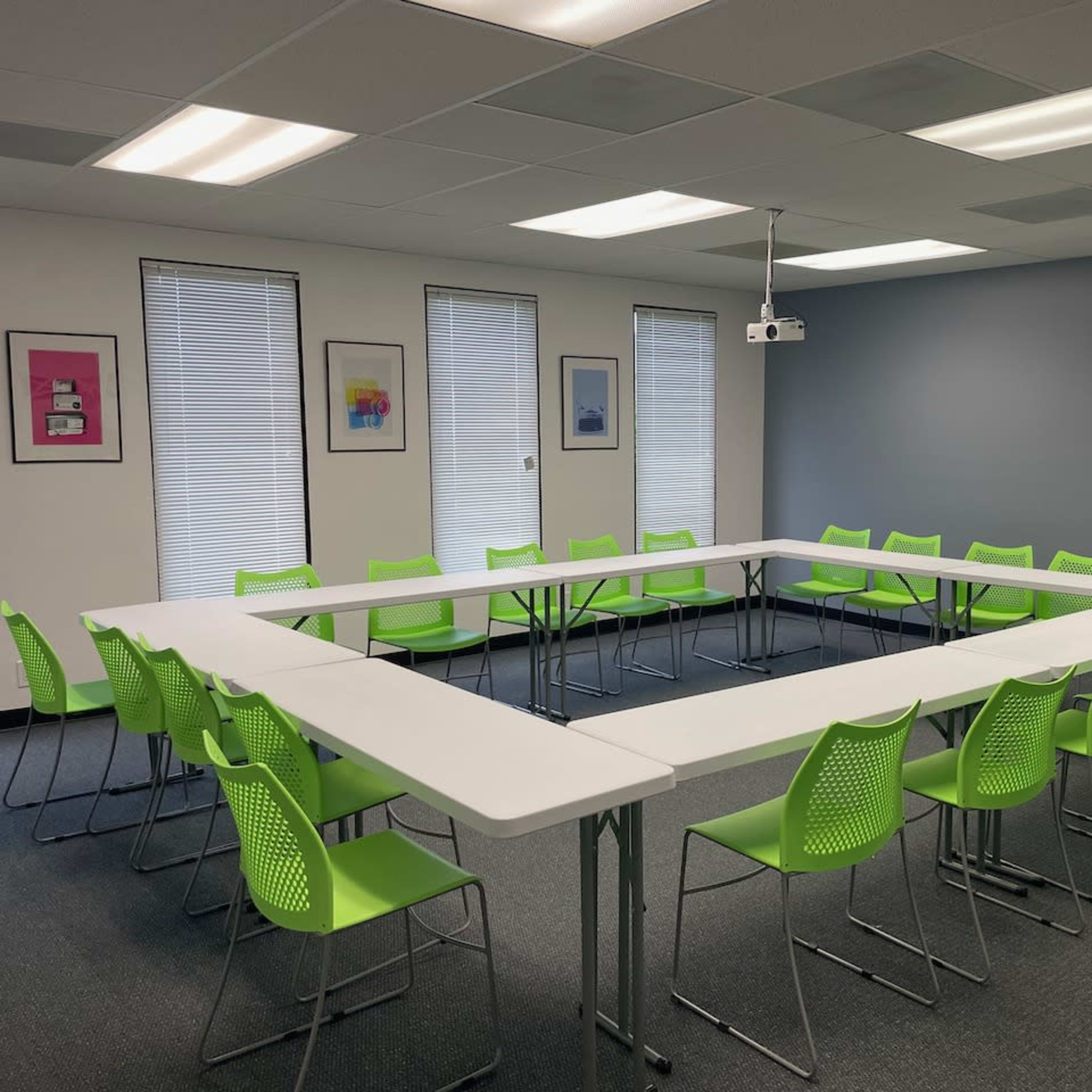 A conference room features a triangular arrangement of white tables surrounded by green chairs, with windows and artwork on the walls.