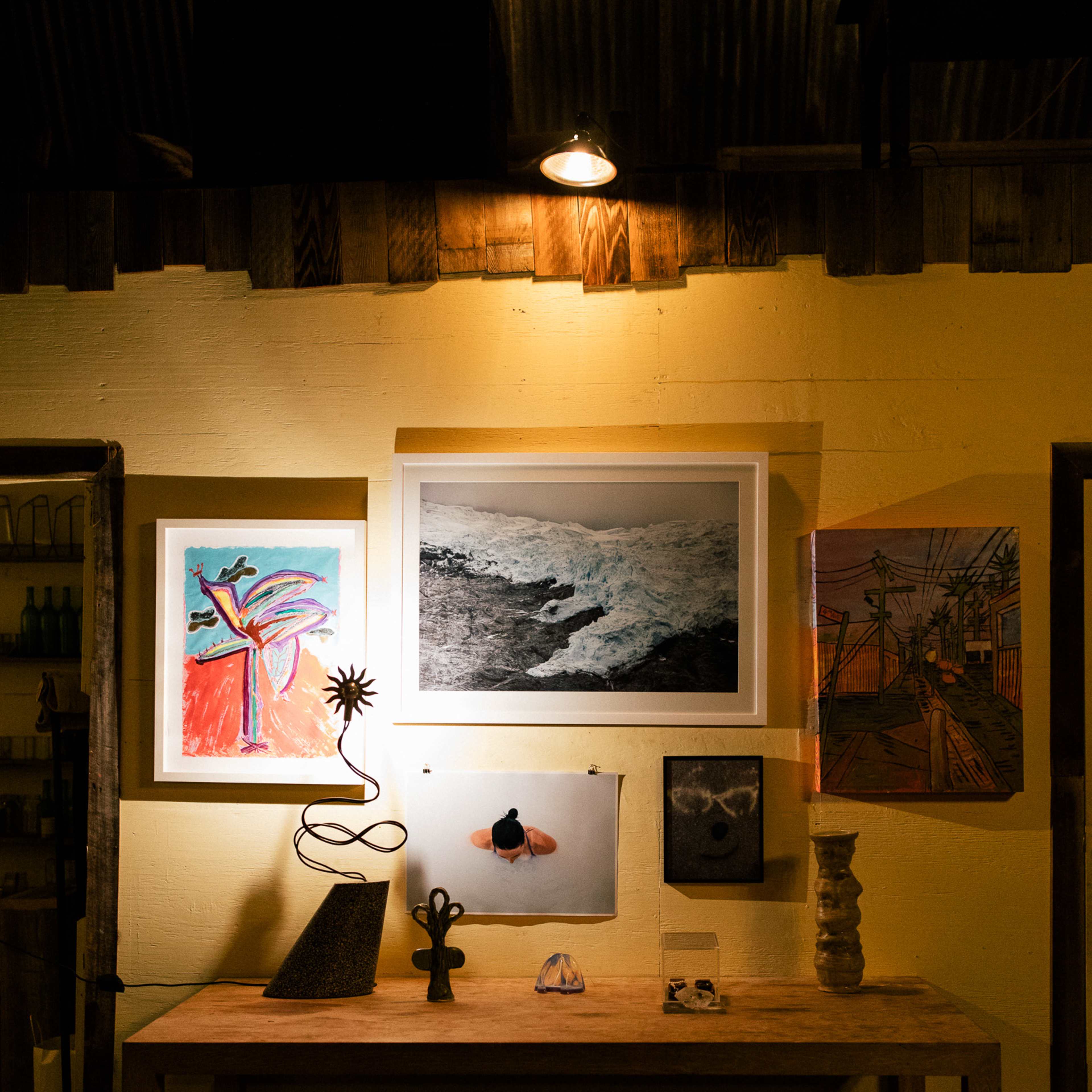 A wooden table displays various artworks and sculptures against a brightly colored wall illuminated by overhead lights.