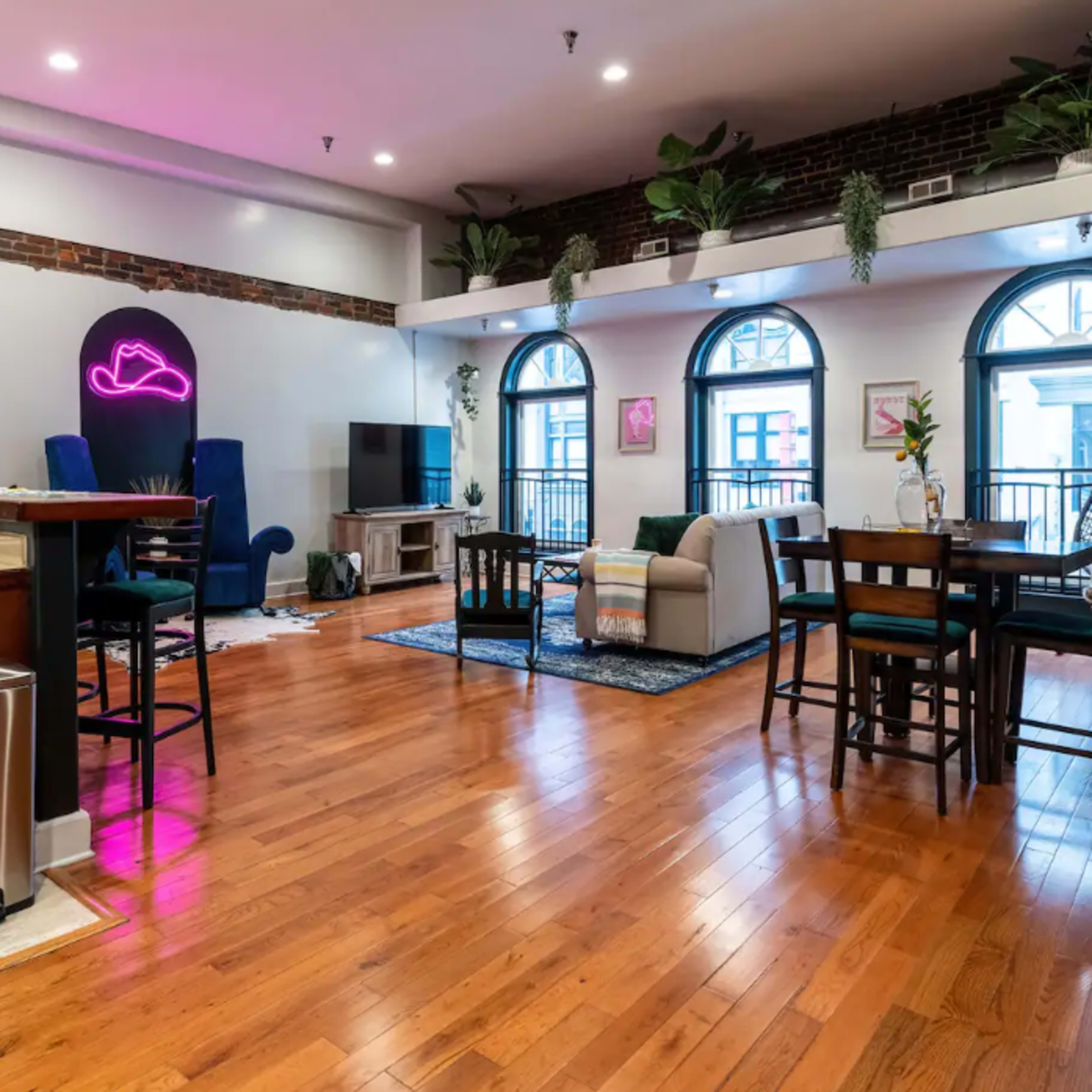 A spacious living area with hardwood floors, a kitchen island, seating arrangements, and large windows illuminated by indoor plants and ambient lighting.