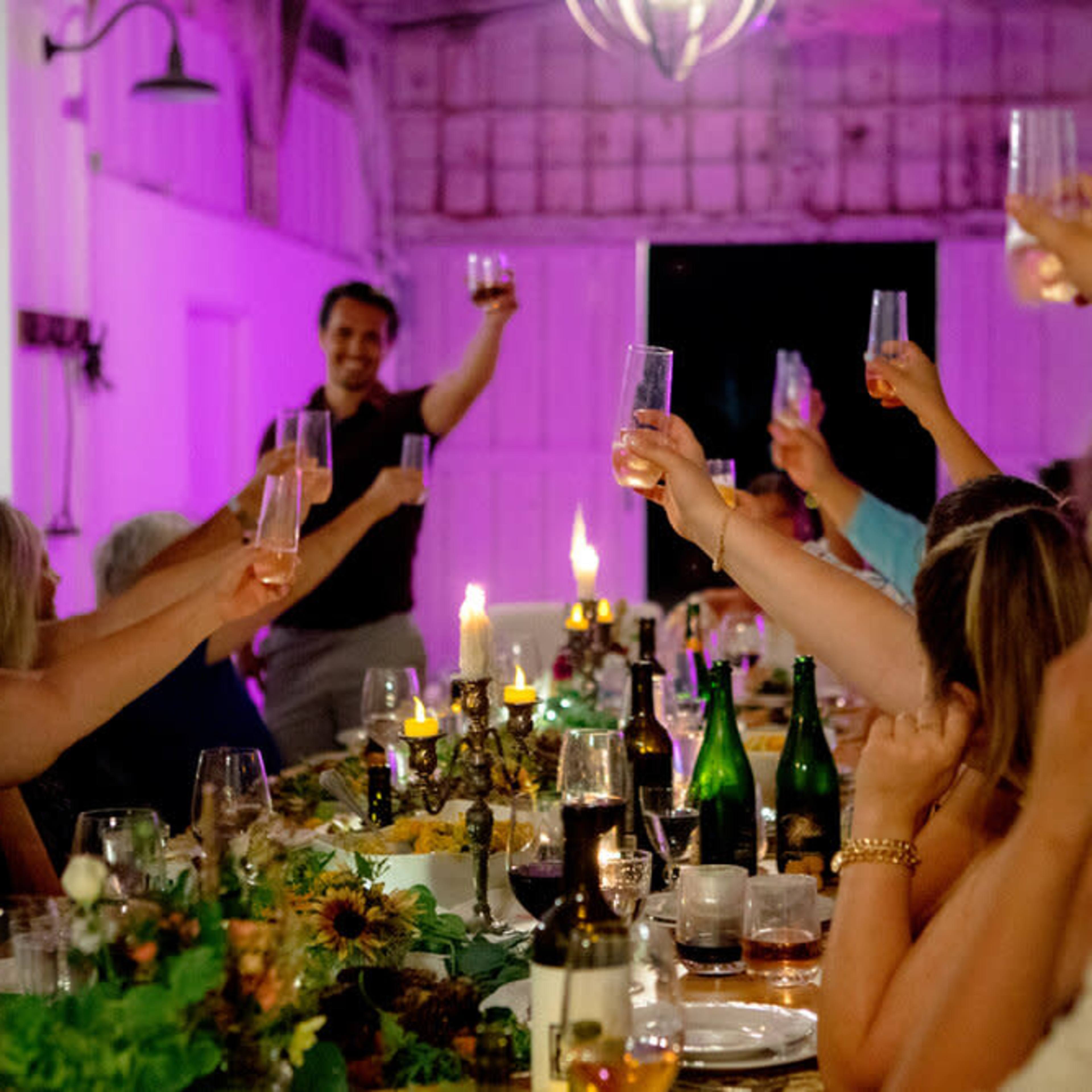 A group of people raises their glasses in a toast at a festive dinner table illuminated by candles and colored lights.