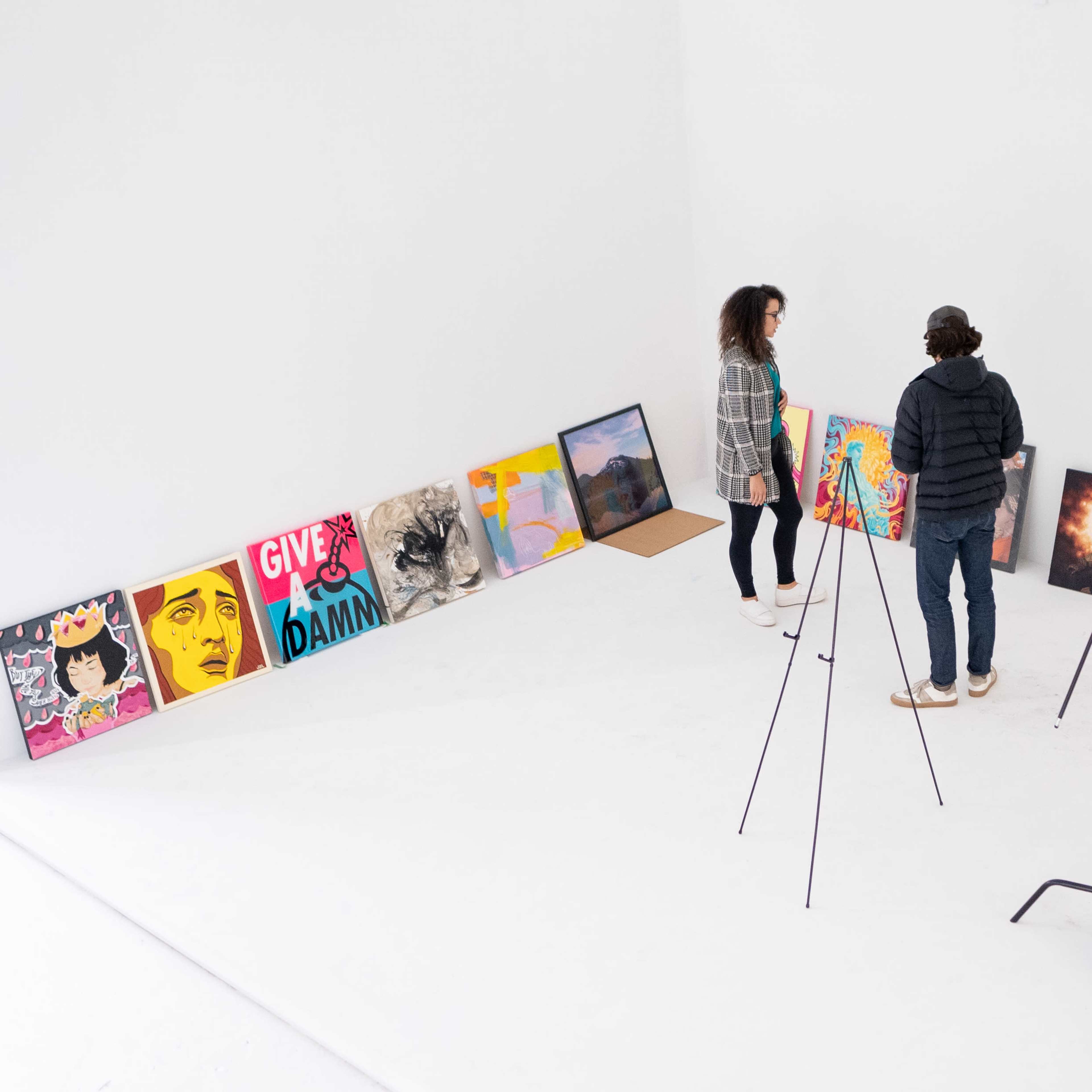 Two people are viewing and discussing a series of colorful artworks displayed along a white wall in a minimally furnished gallery space, with a large softbox light set up nearby.