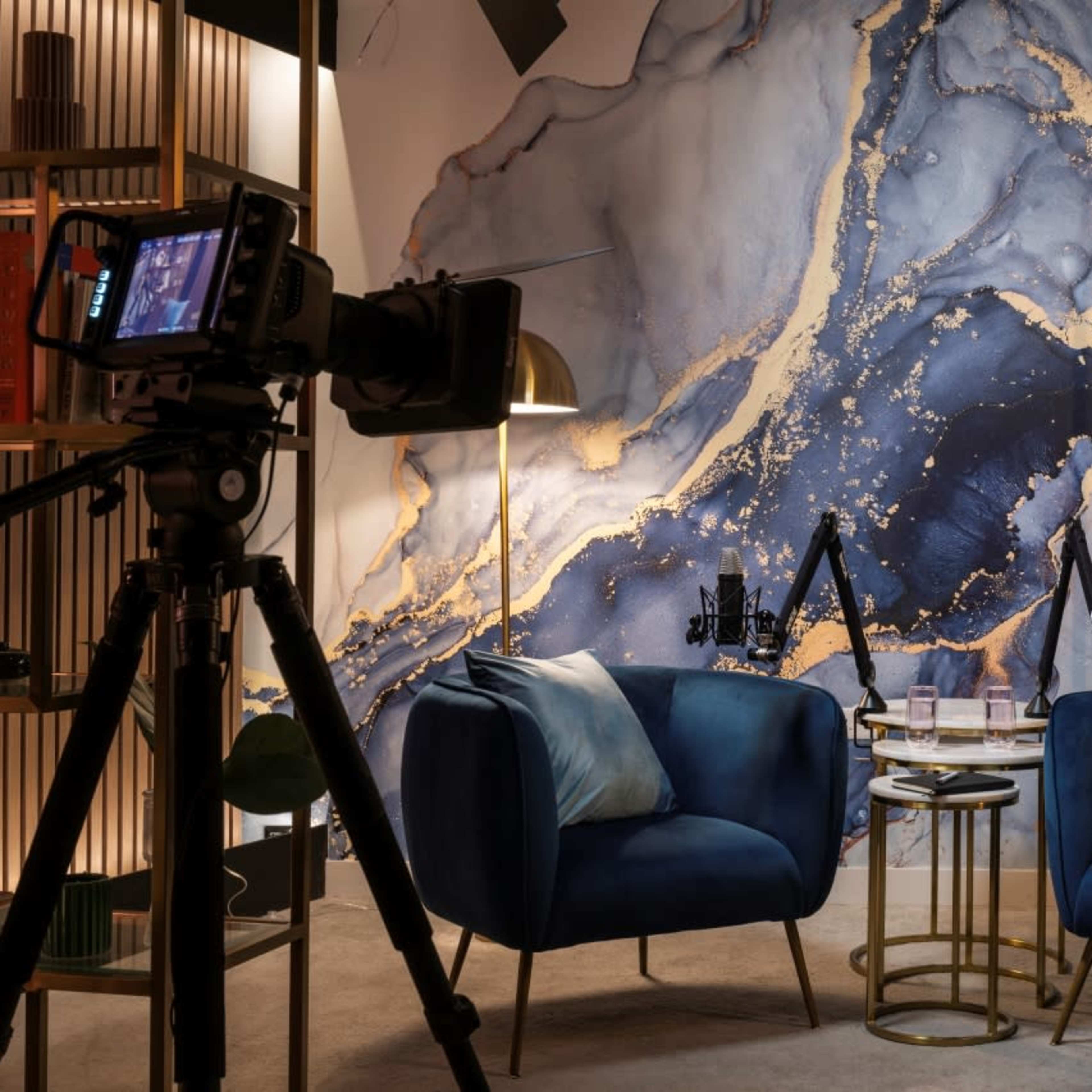A modern podcast studio with two blue chairs facing a small table, surrounded by cameras and a decorative wall featuring abstract patterns.