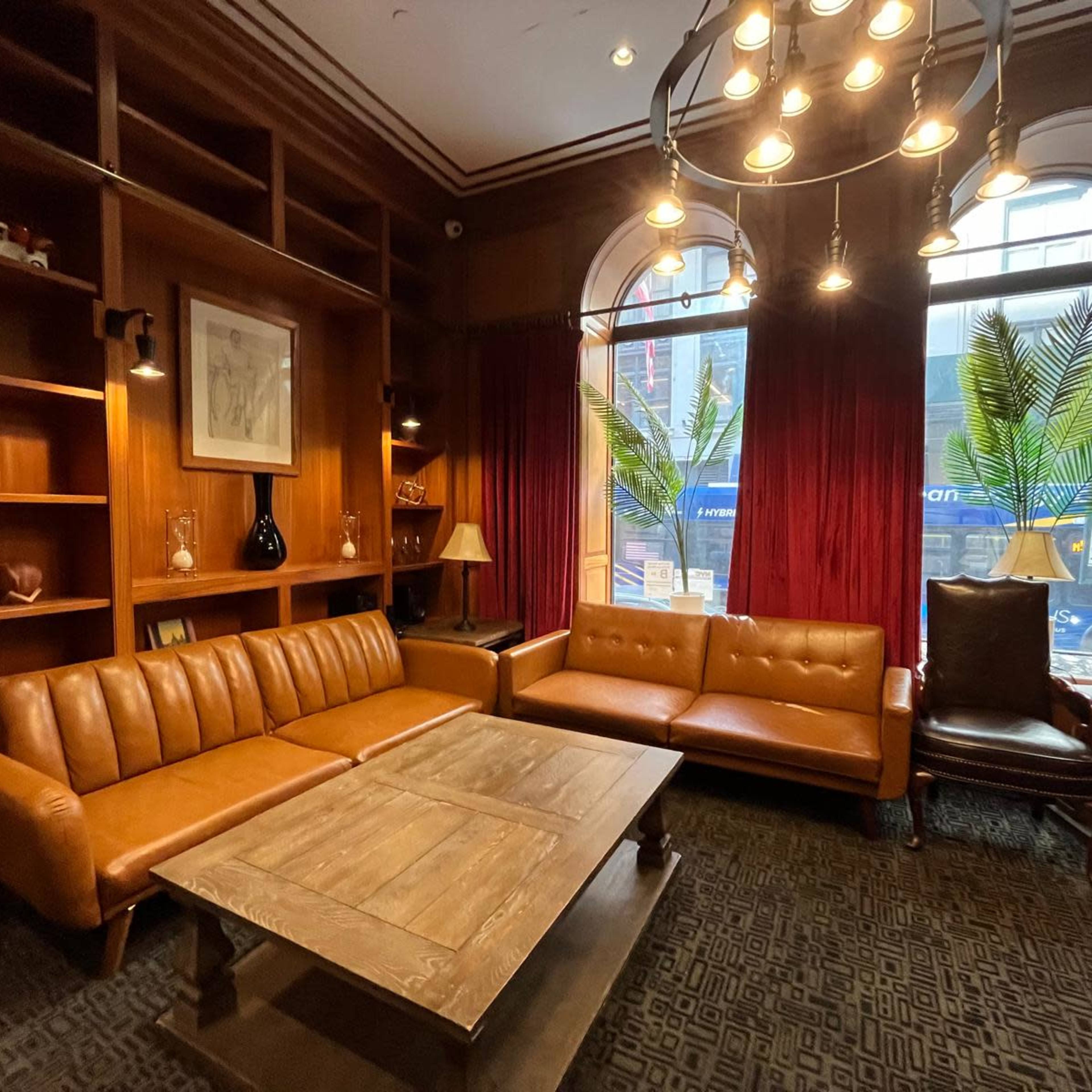 A cozy lounge area with brown leather sofas, a wooden coffee table, and large windows featuring red curtains.