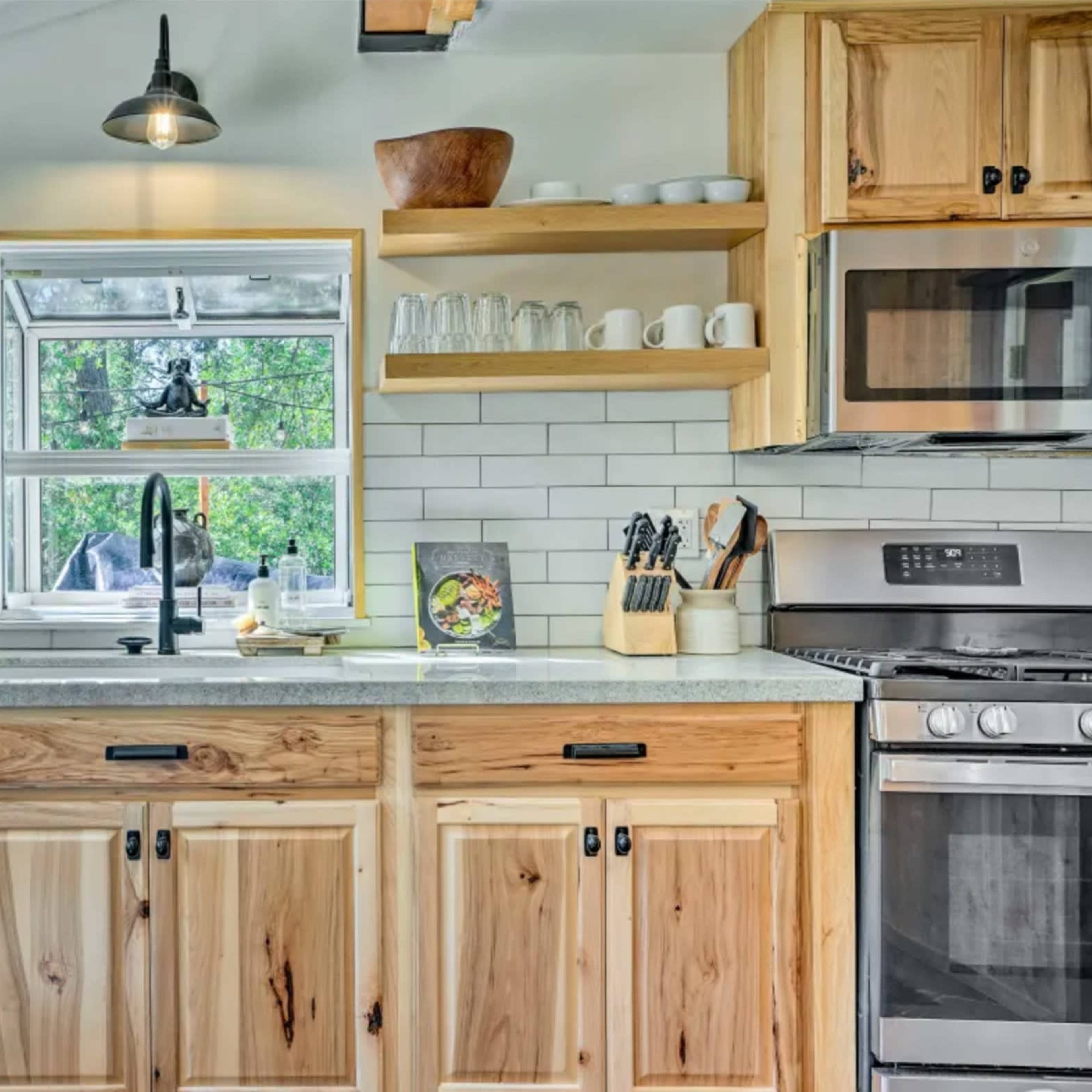 A modern kitchen with wooden cabinets, open shelving above a sink, stainless steel appliances, and a window providing natural light.