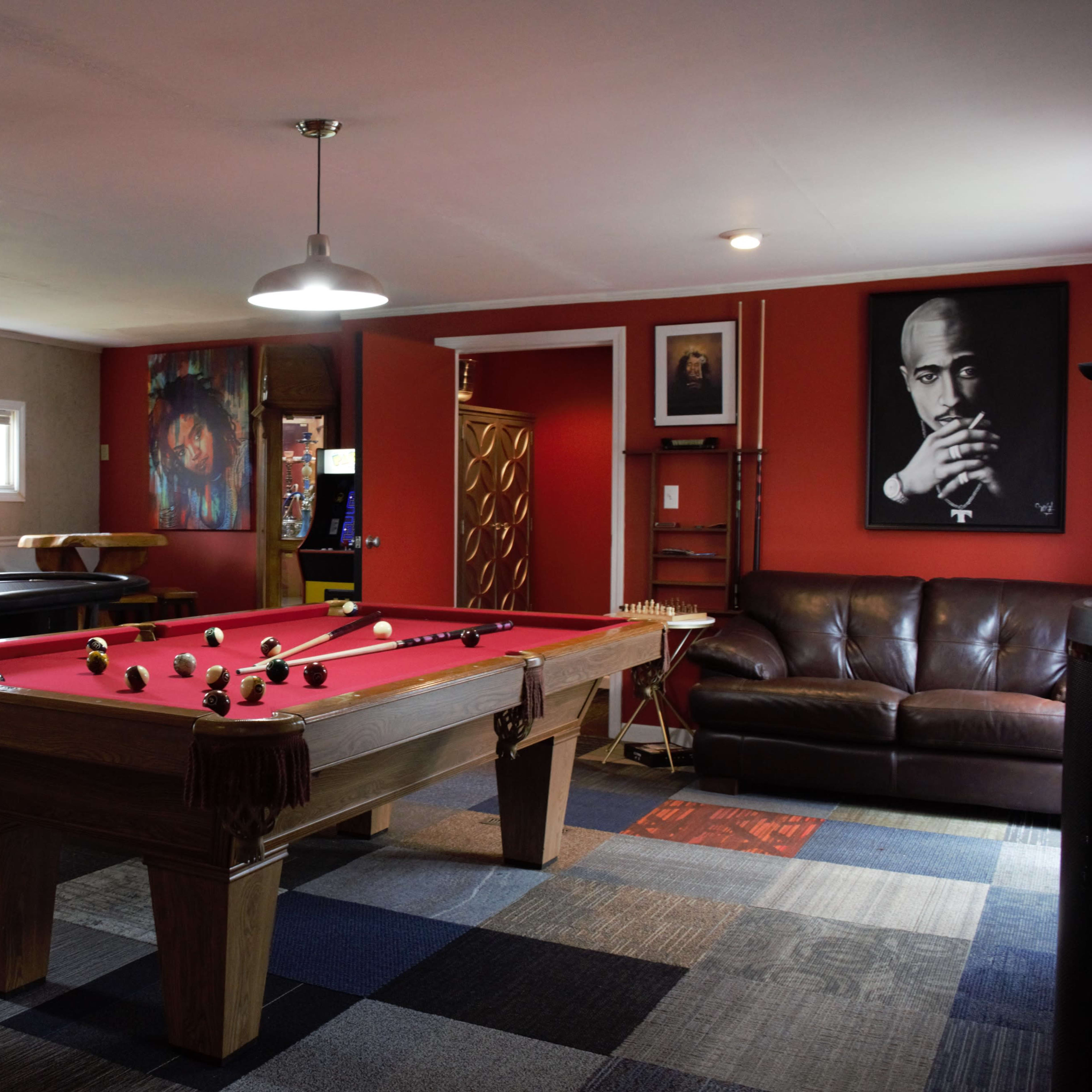 A game room featuring a red-felted pool table, a dark couch, wall art of notable figures, and a flat-screen TV on one wall.
