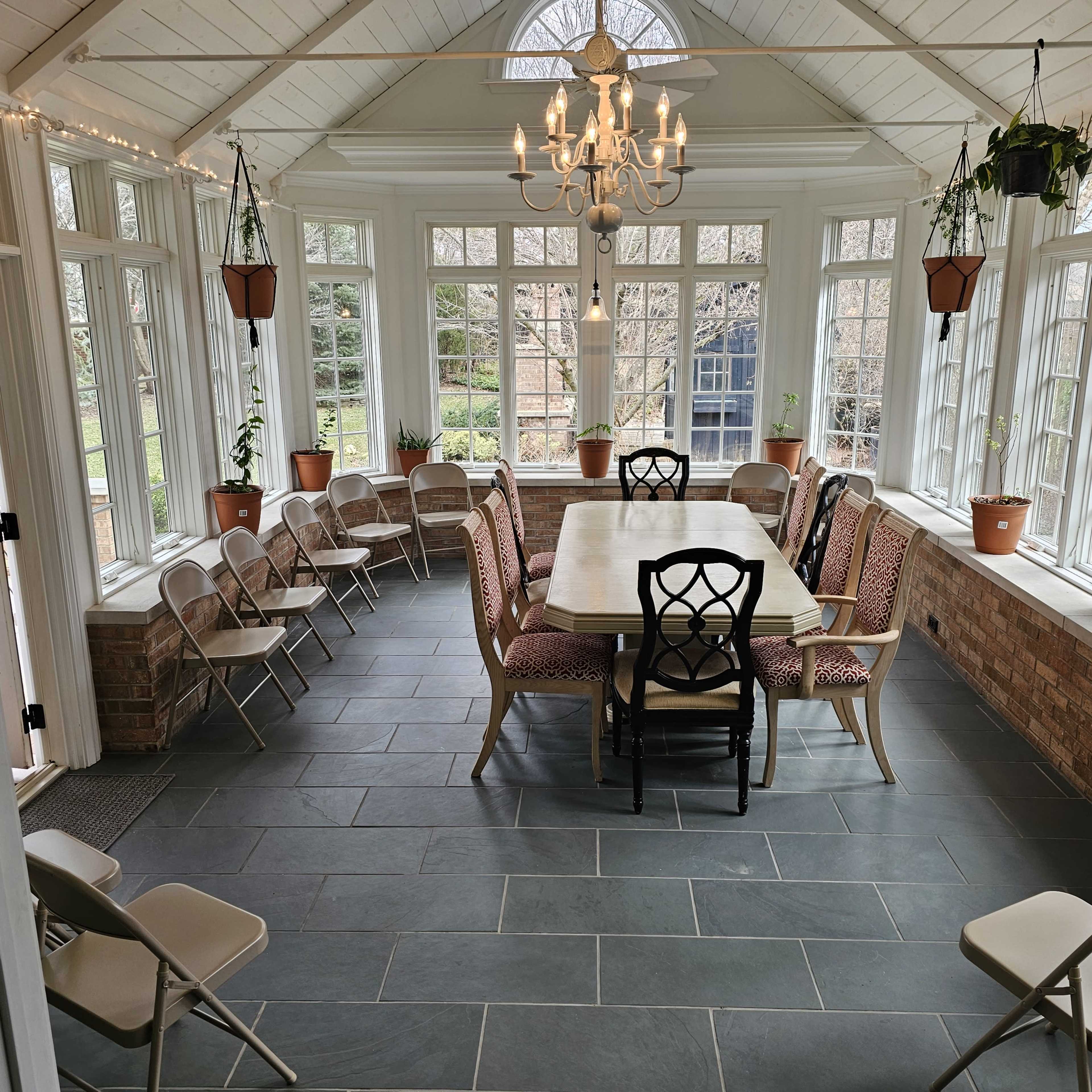 A sunlit conservatory features a large table surrounded by chairs and potted plants, with large windows providing a view of the garden.