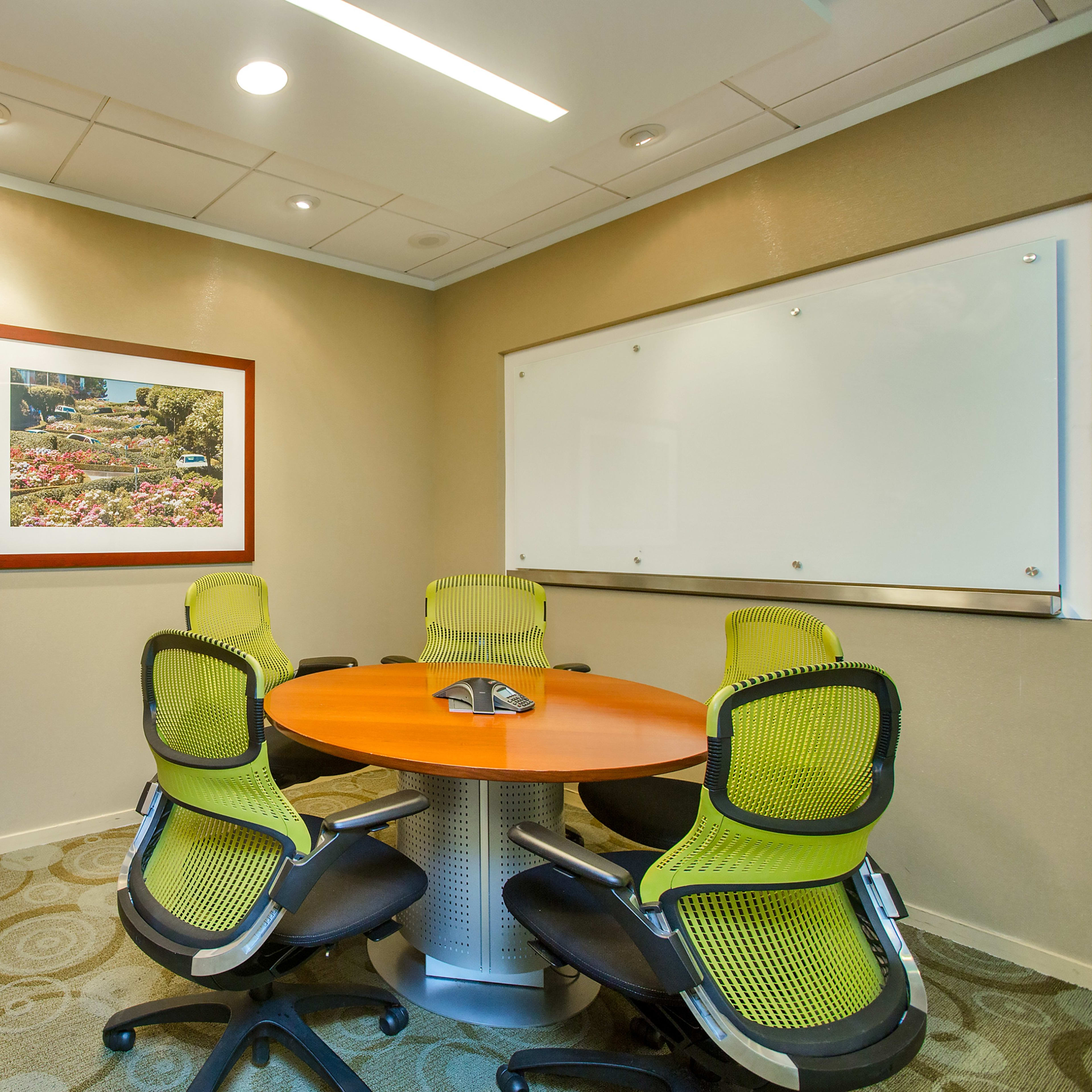 A small meeting room features a circular table surrounded by six green chairs, a framed landscape photograph on the wall, and a whiteboard.