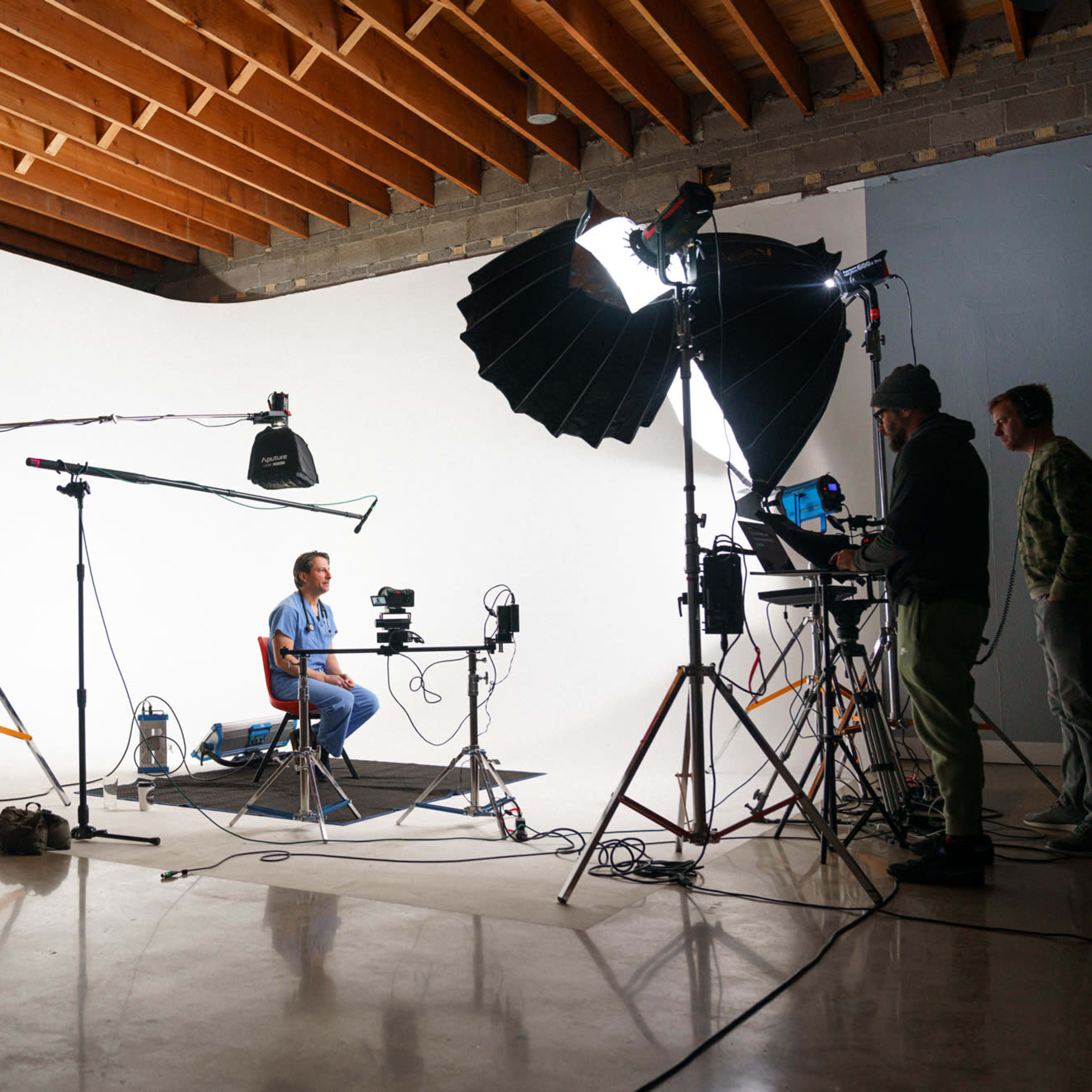 A man sits at a table in front of a white backdrop while a camera and lighting equipment are set up nearby.