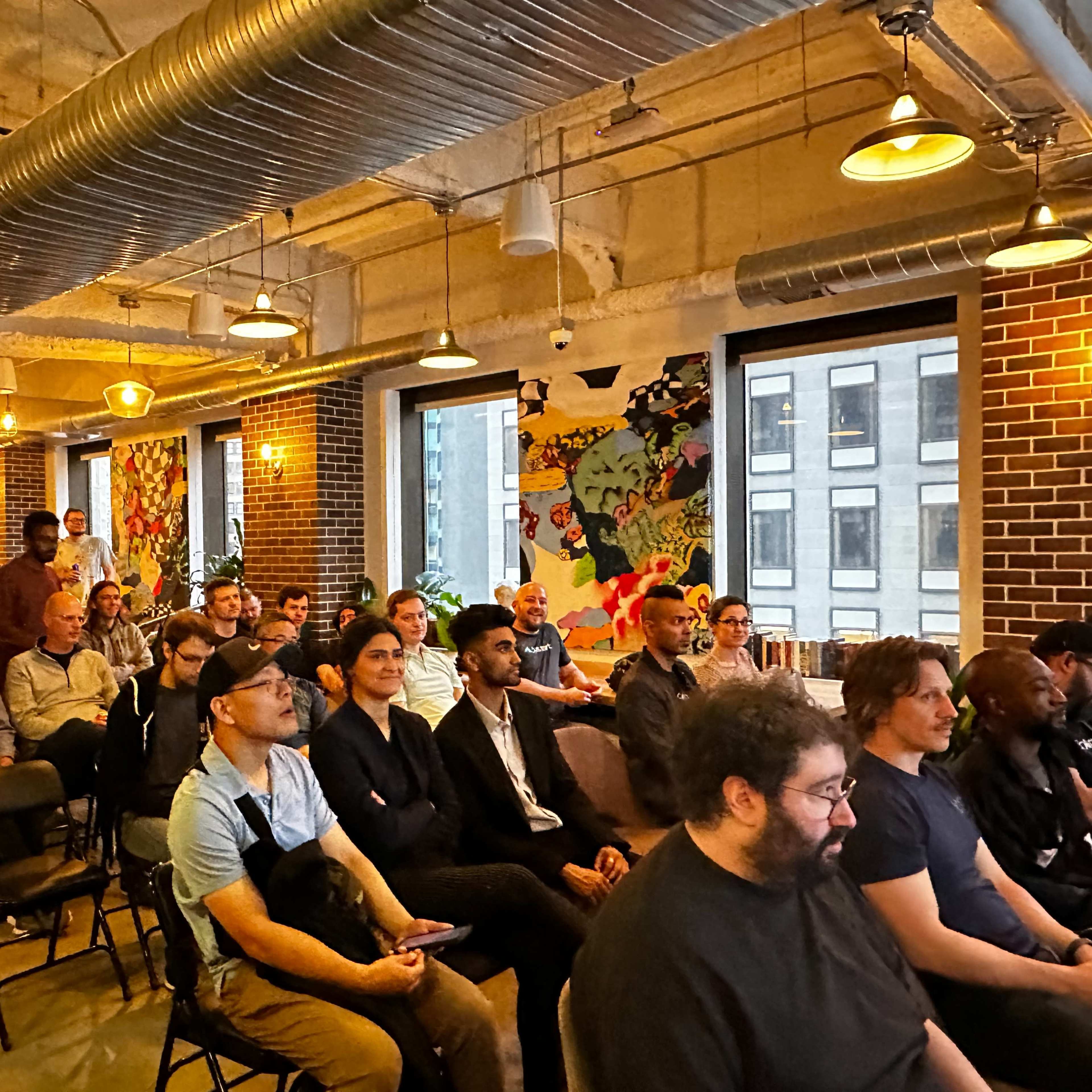 A diverse group of people sits in rows of chairs, facing a speaker at a brightly lit event space with colorful wall art.