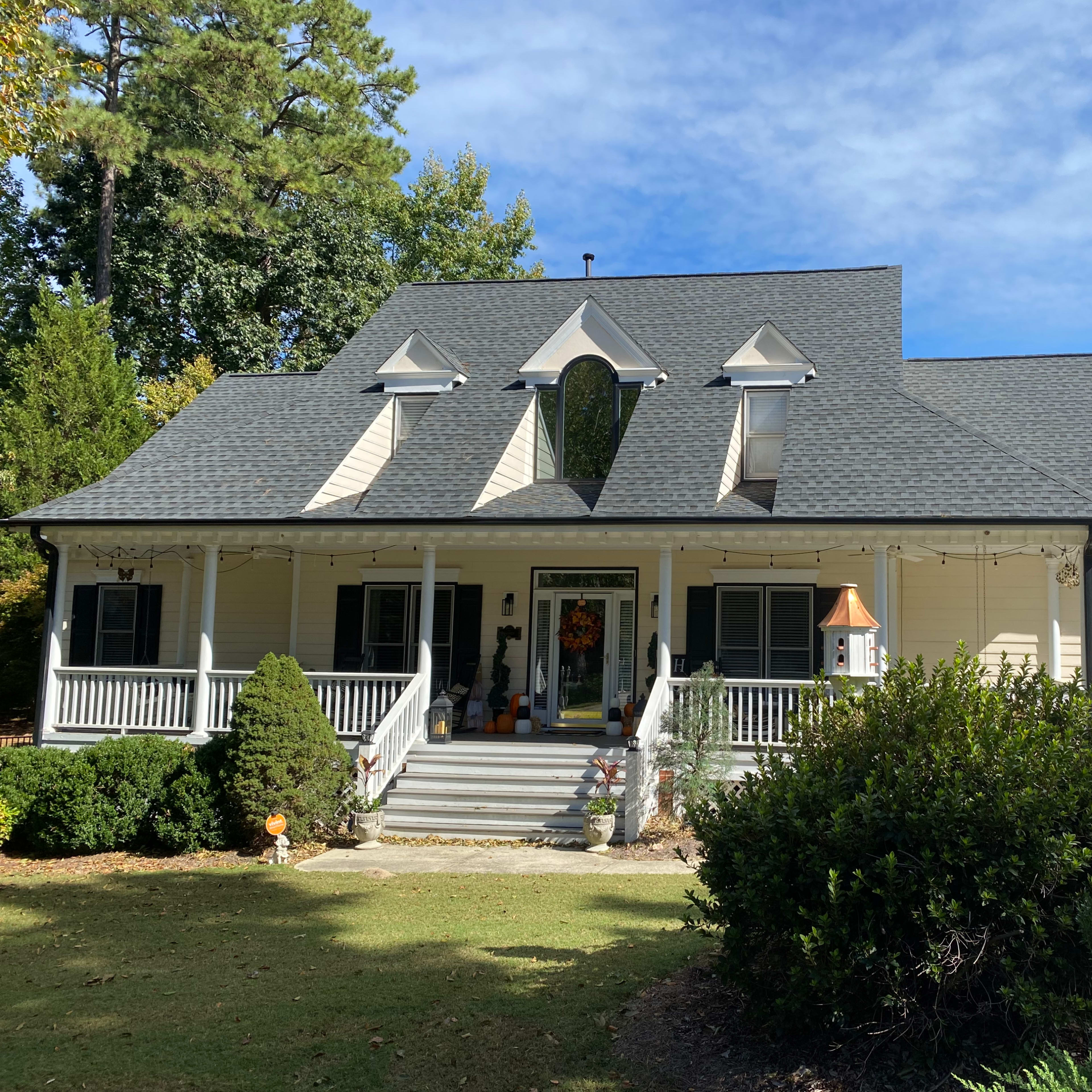 A large, light-colored house with a steep roof and a front porch is surrounded by green shrubs and trees.