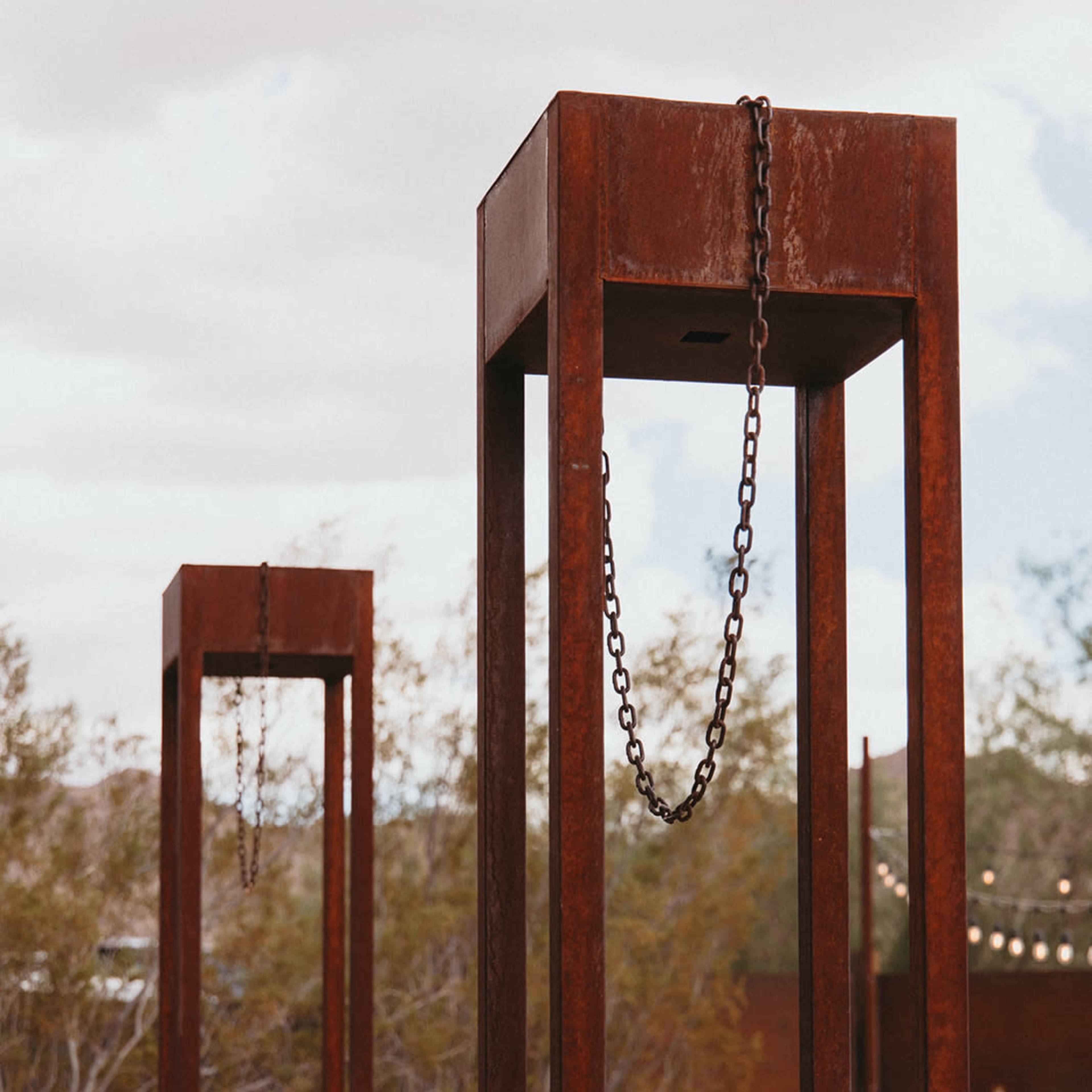 Two tall, rust-colored metal structures with chains hanging from their tops, set against a desert landscape filled with plants and rocks.