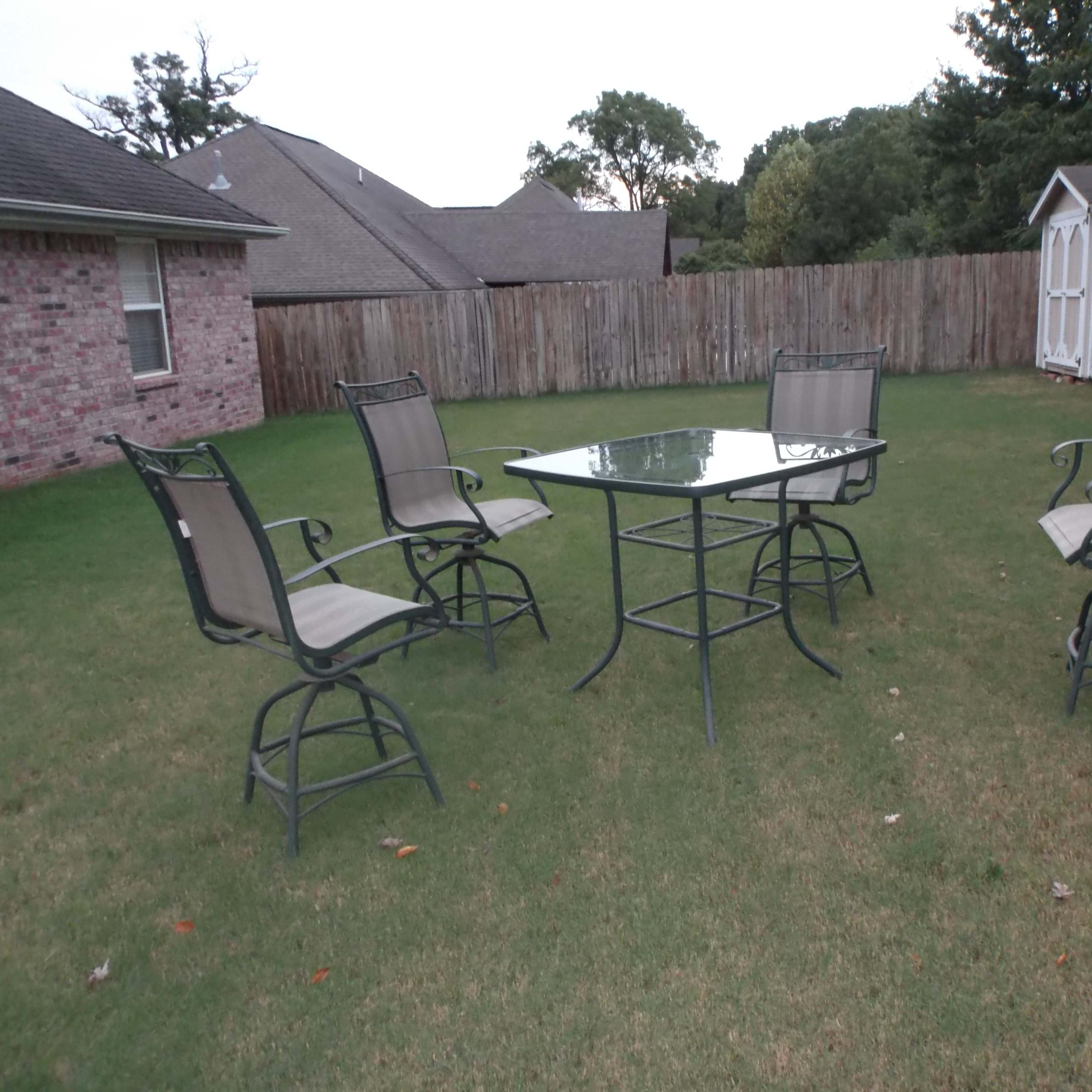 A set of four metal chairs surrounds a glass-topped table on a grassy lawn, with a wooden fence and storage shed visible in the background.