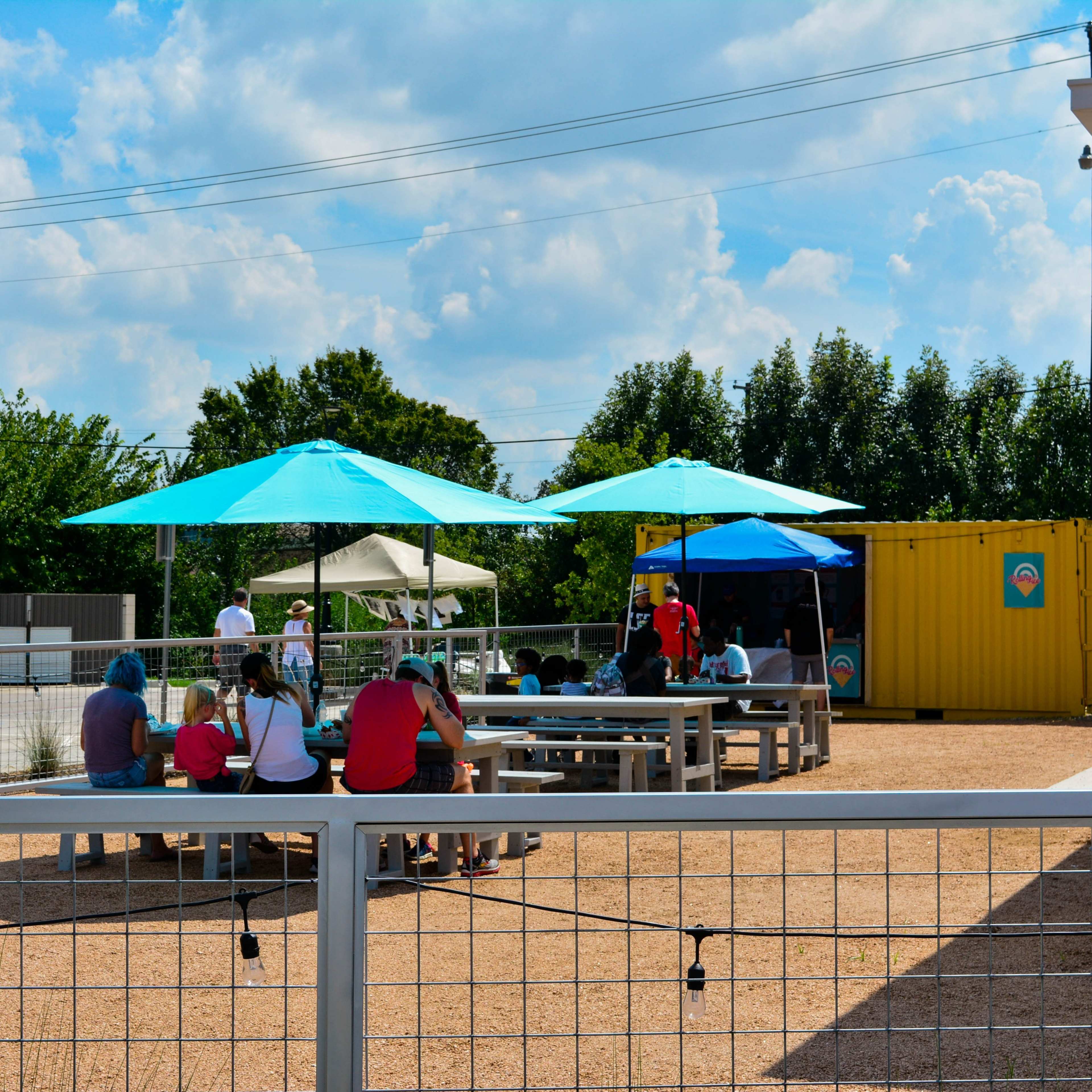 A group of people sit at tables under blue and white umbrellas in an outdoor area near a yellow shipping container.