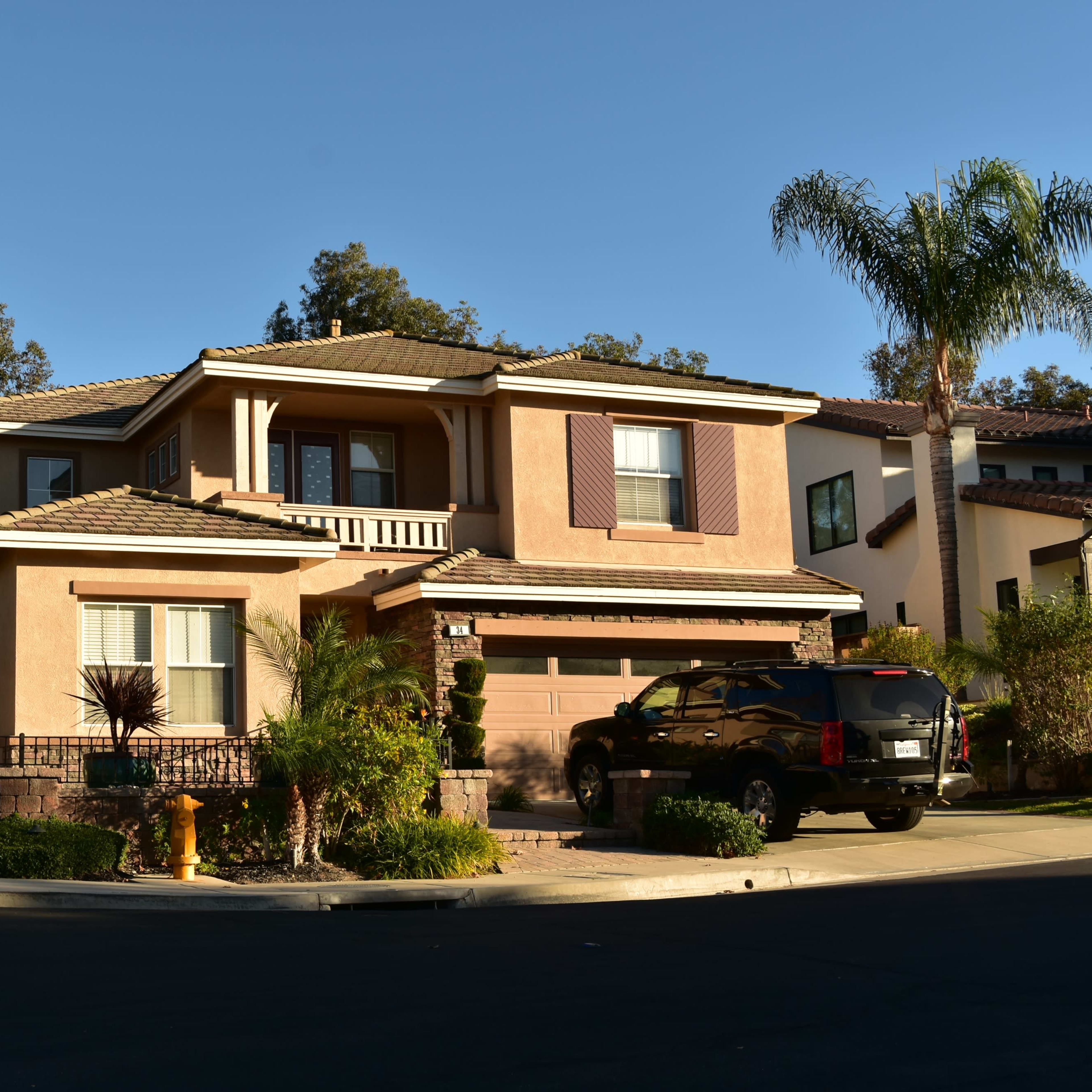A two-story house with a tan exterior, surrounded by greenery and a palm tree, located on a corner lot in a suburban neighborhood.