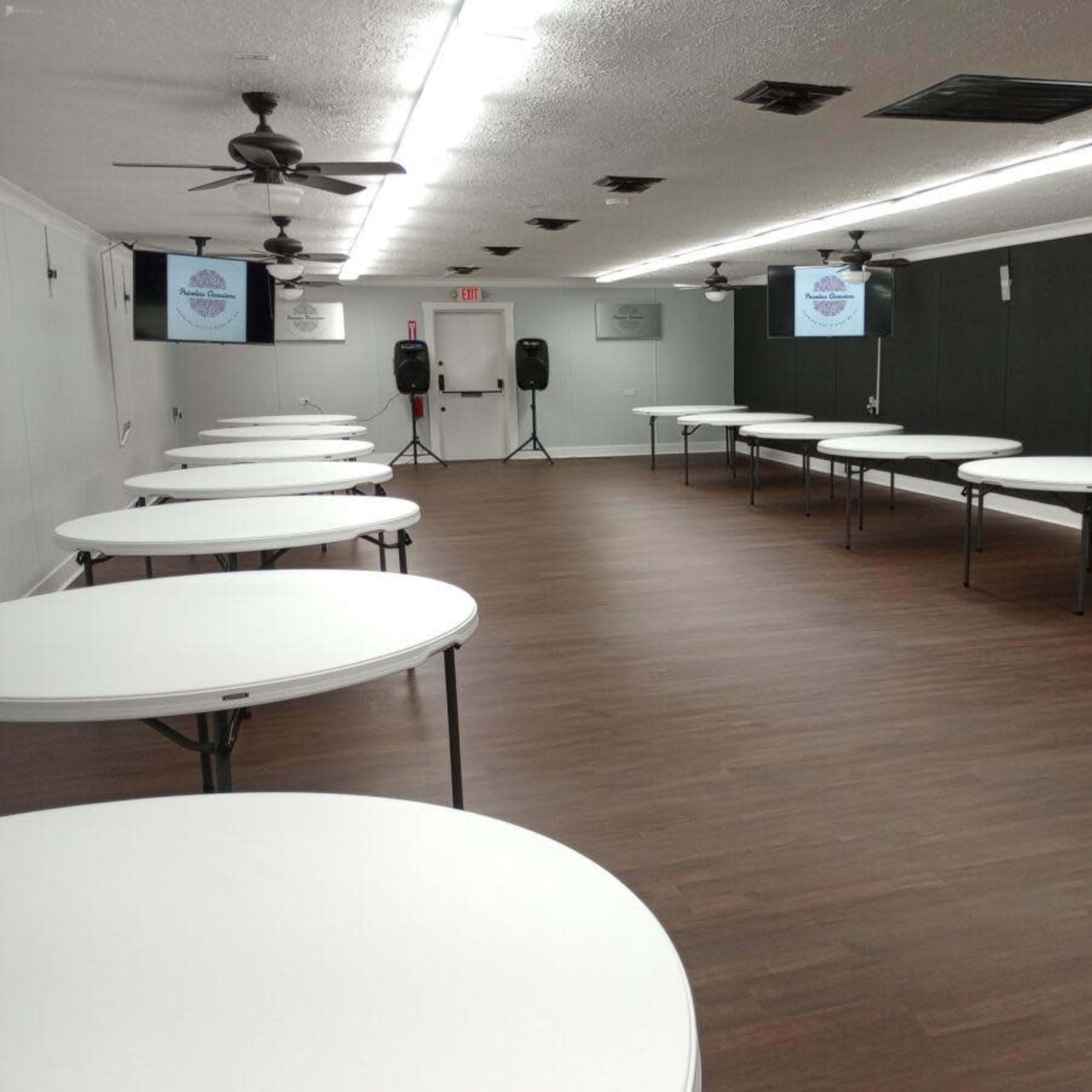 A spacious room set up with several round tables arranged for an event, featuring a wooden floor and ceiling fans.