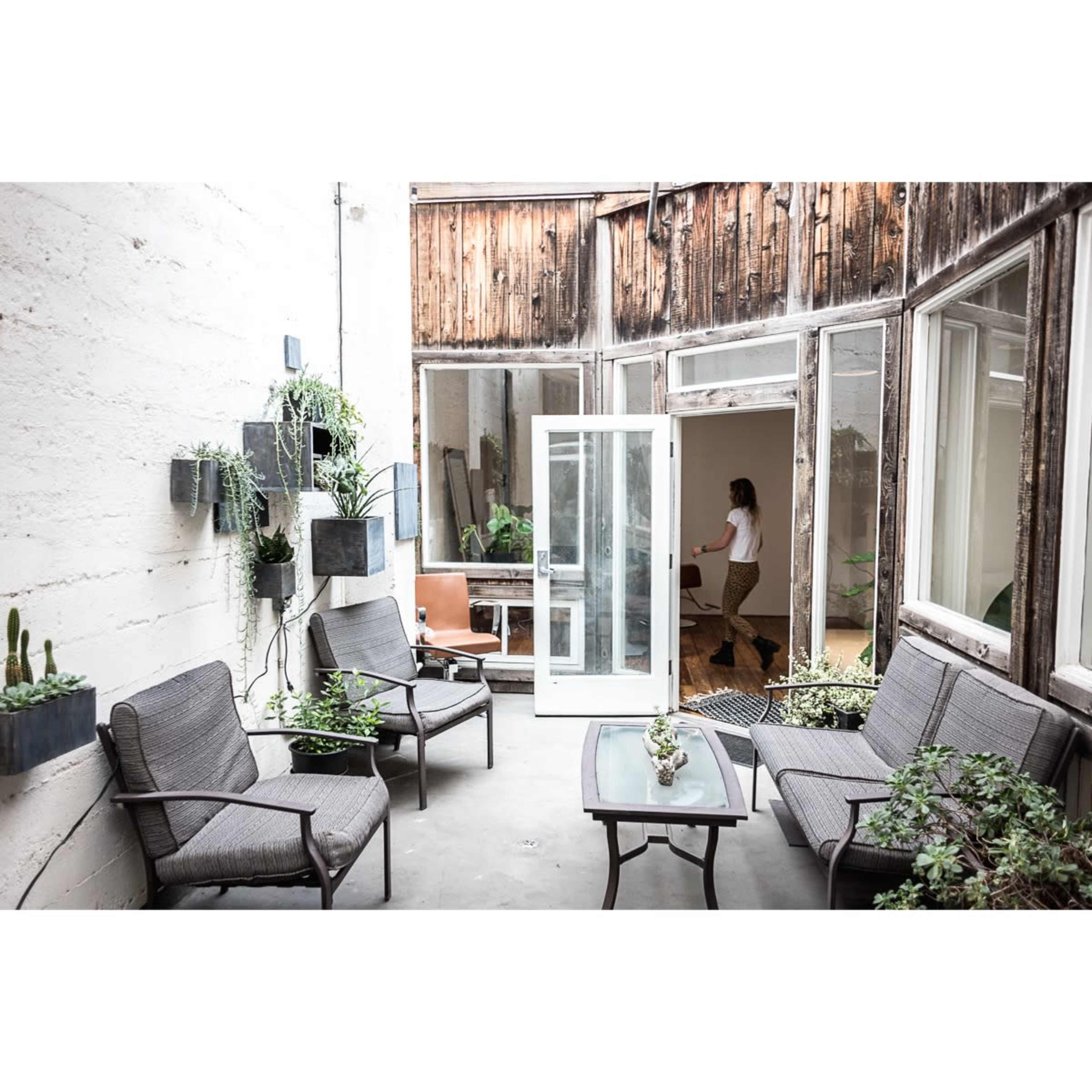 A small, modern courtyard features potted plants, metal furniture, and large windows, with a figure standing inside an adjoining room.