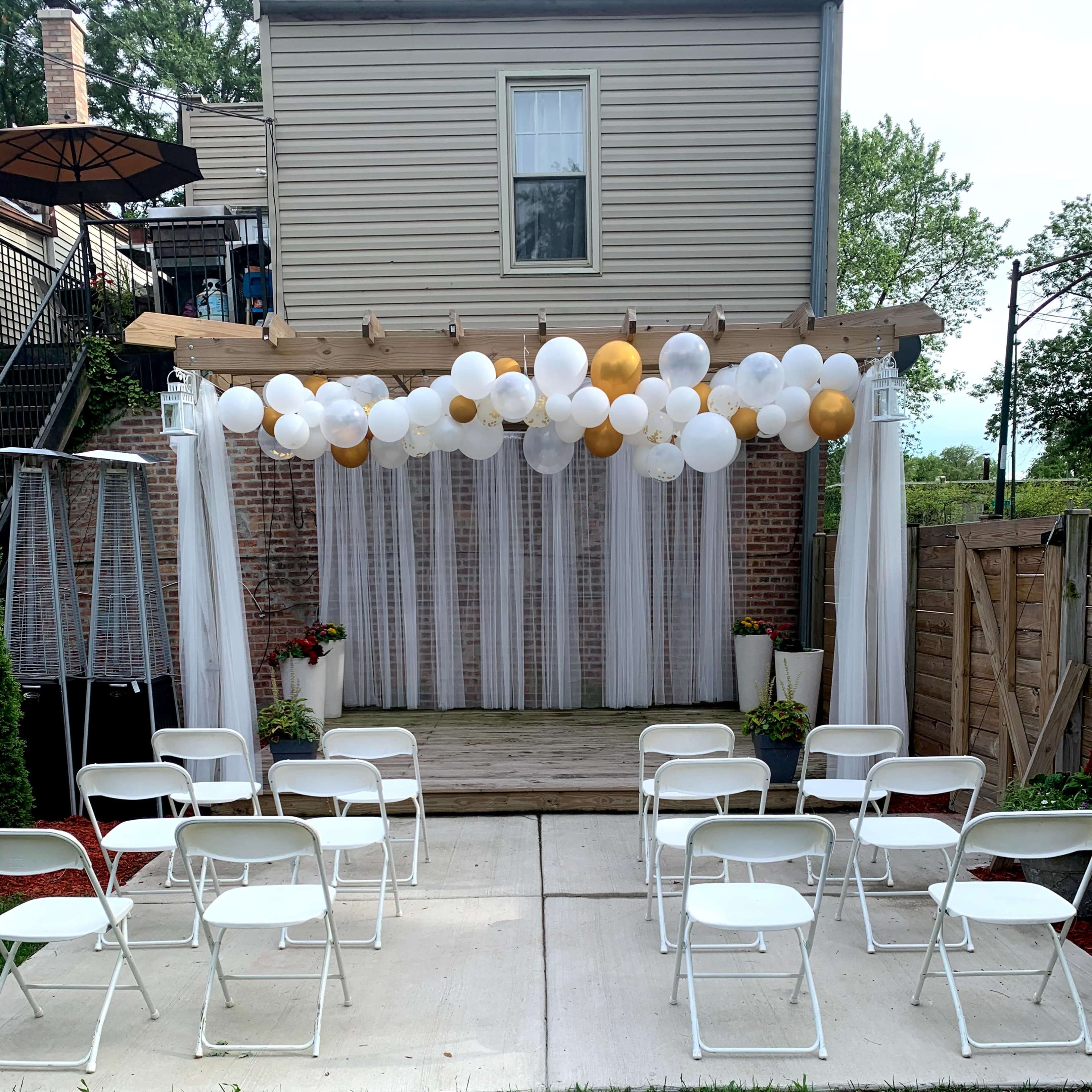 A decorated outdoor stage area with white chairs arranged in front of a wooden platform, draped with white fabric and adorned with balloons in various colors.