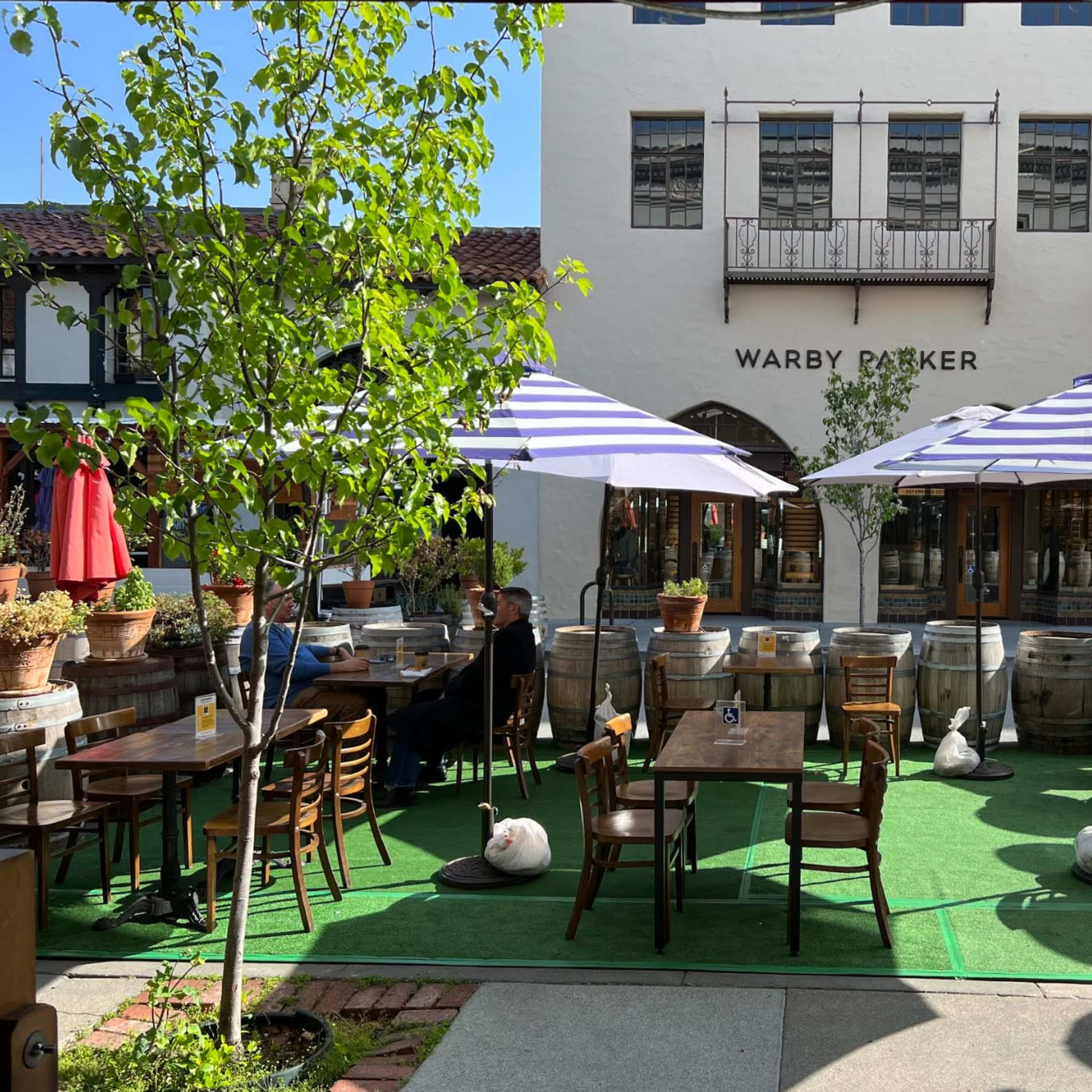 An outdoor dining area with tables and chairs under striped umbrellas, surrounded by potted plants and barrels near a storefront labeled "WARBY PARKER."