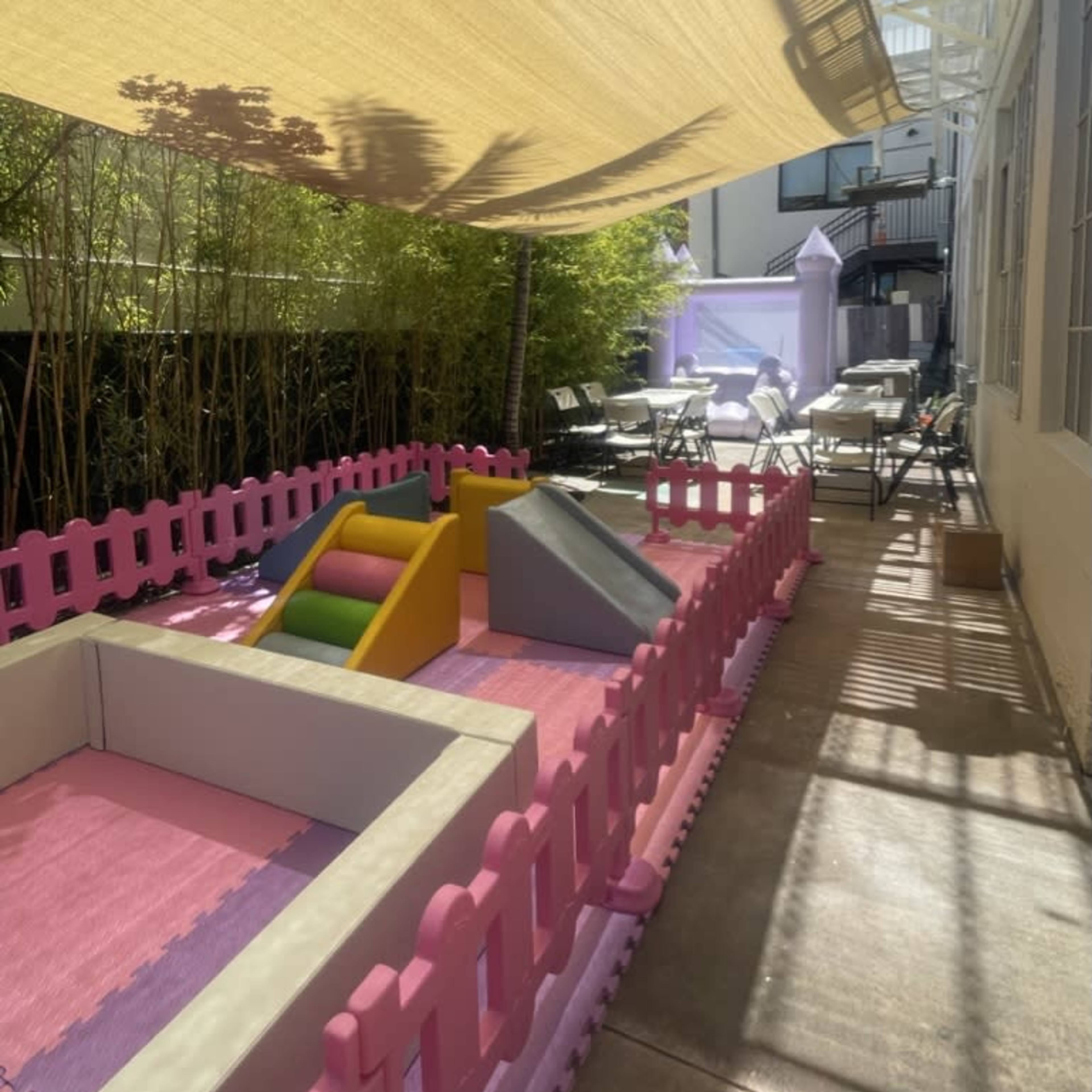A shaded outdoor play area enclosed by a pink fence, featuring a small slide and climbing structure on soft flooring.