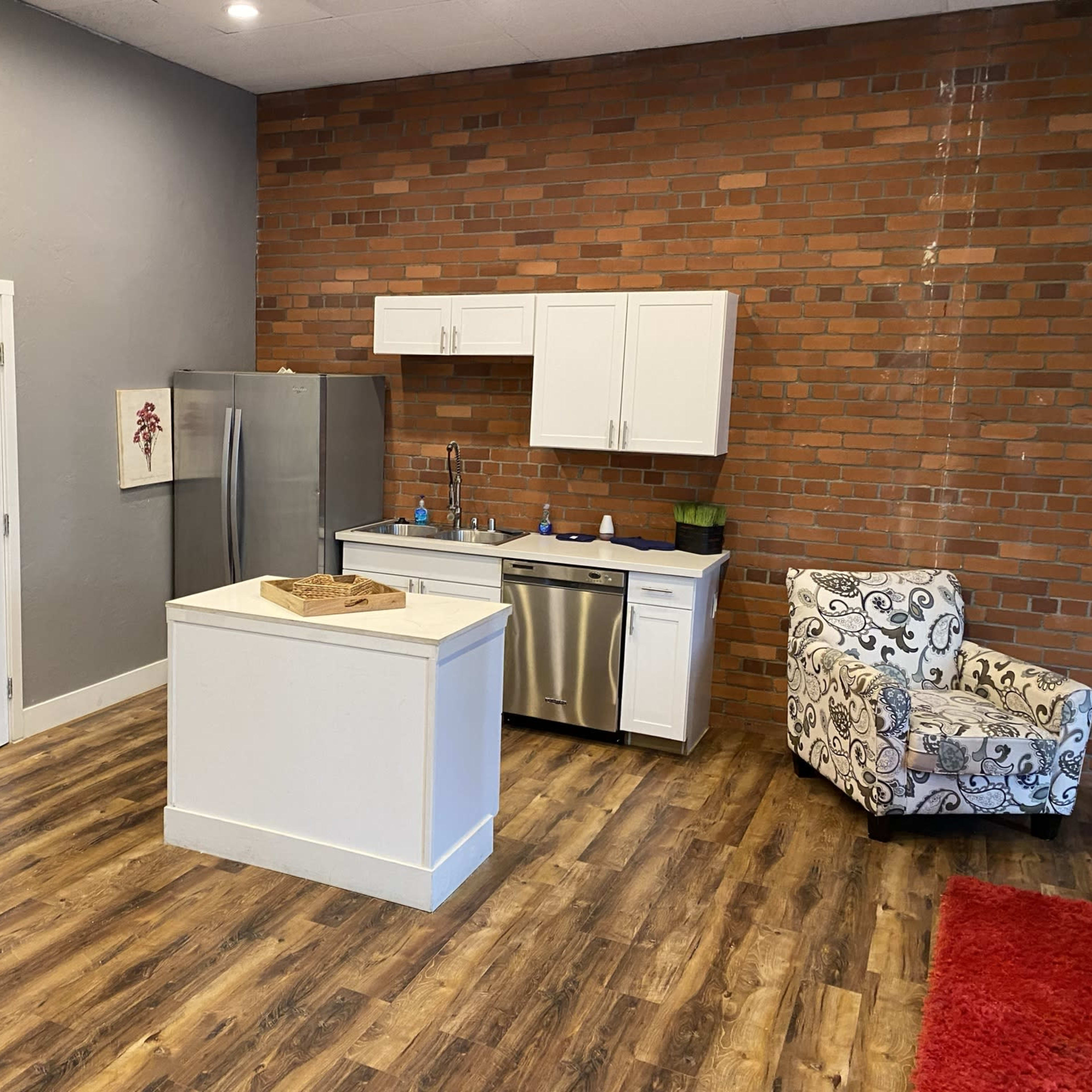 A modern kitchen area with a stainless steel refrigerator, white cabinets, and a central island, set against a backdrop of exposed brick walls and wooden flooring.