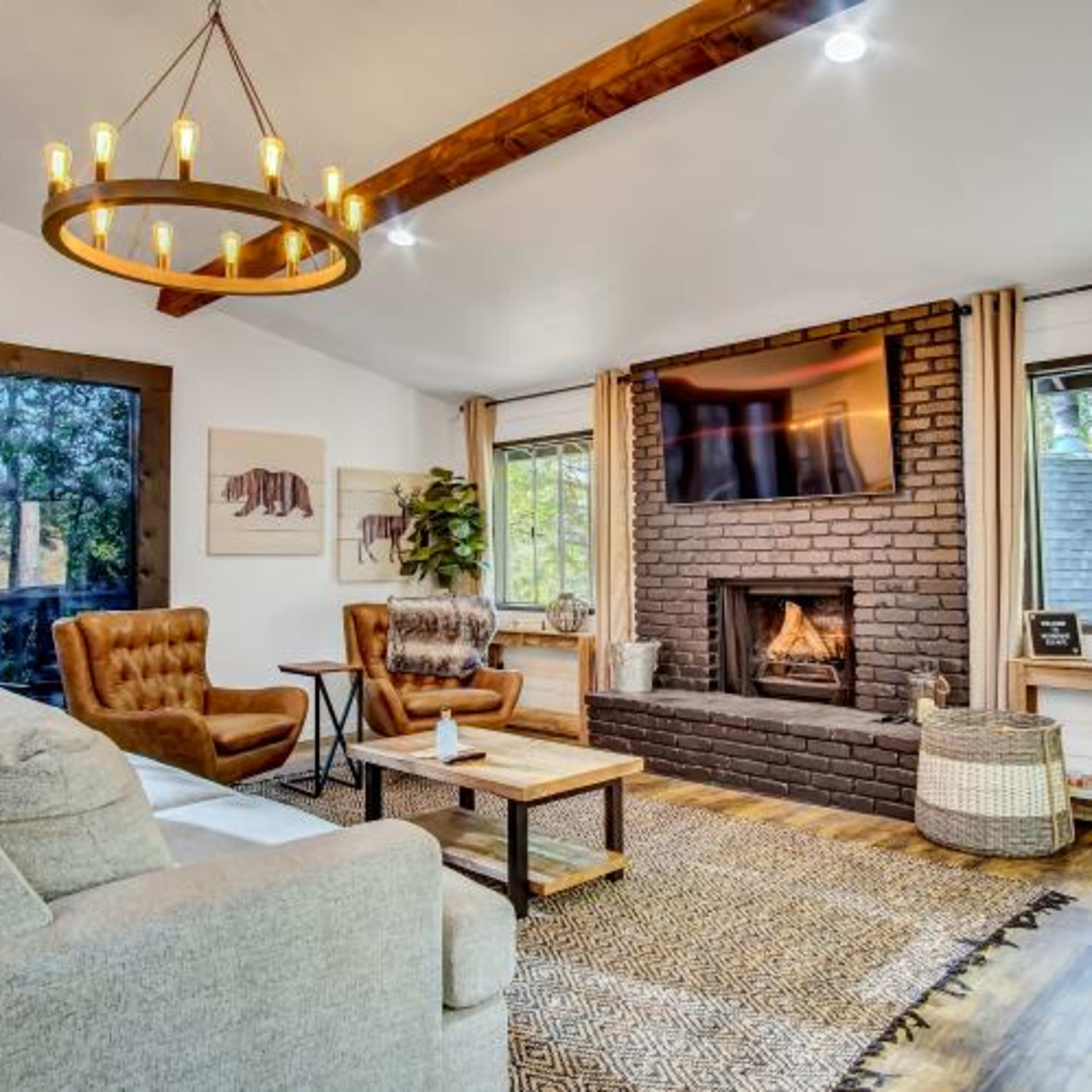 A cozy living room features a fireplace, a flat-screen TV mounted on a black brick wall, and comfortable seating with large windows letting in natural light.