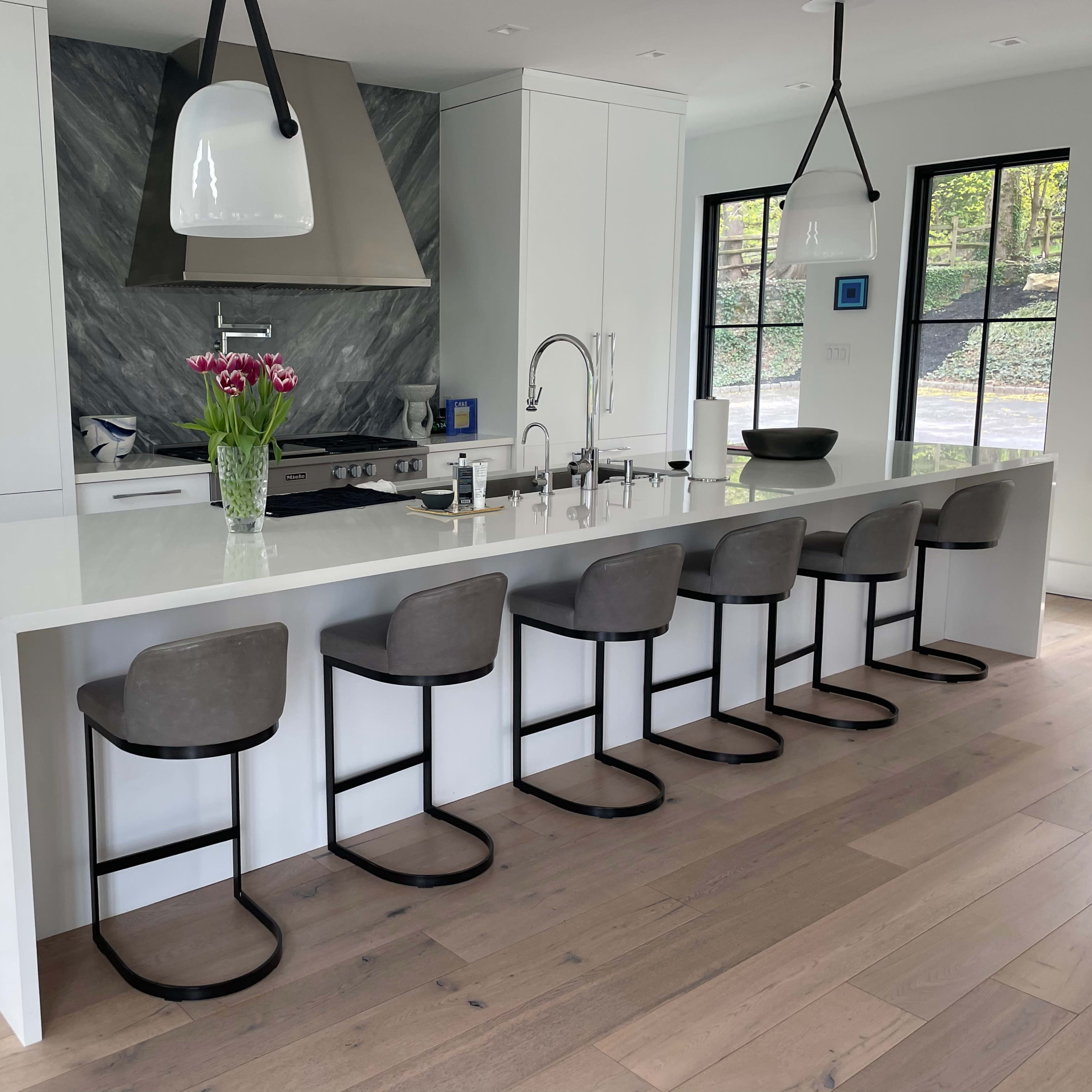 A modern kitchen with a white island countertop, six gray bar stools, and a marble backsplash behind a stainless steel stove.