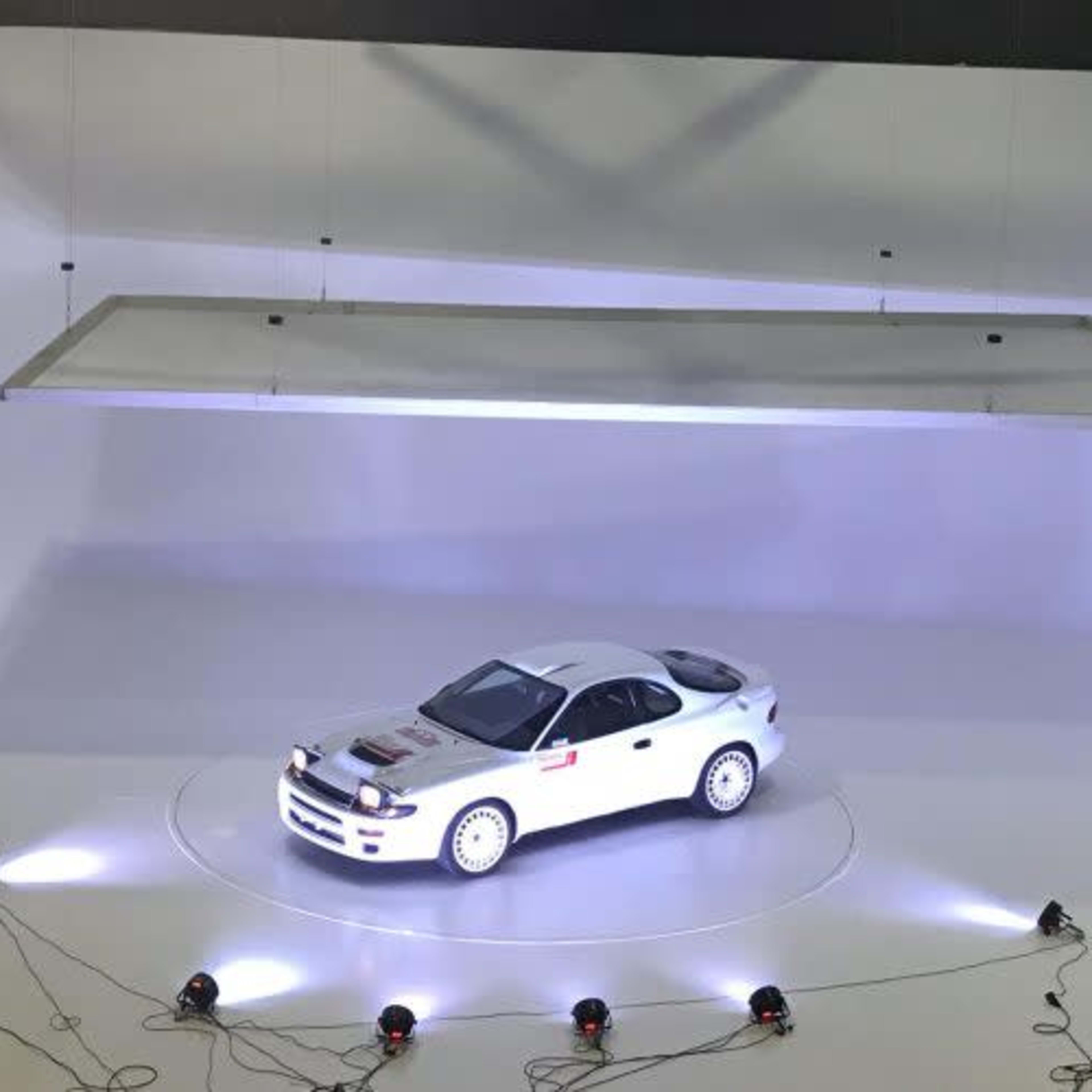 A white sports car is shown on a circular platform under bright lights in a minimalistic studio setting.