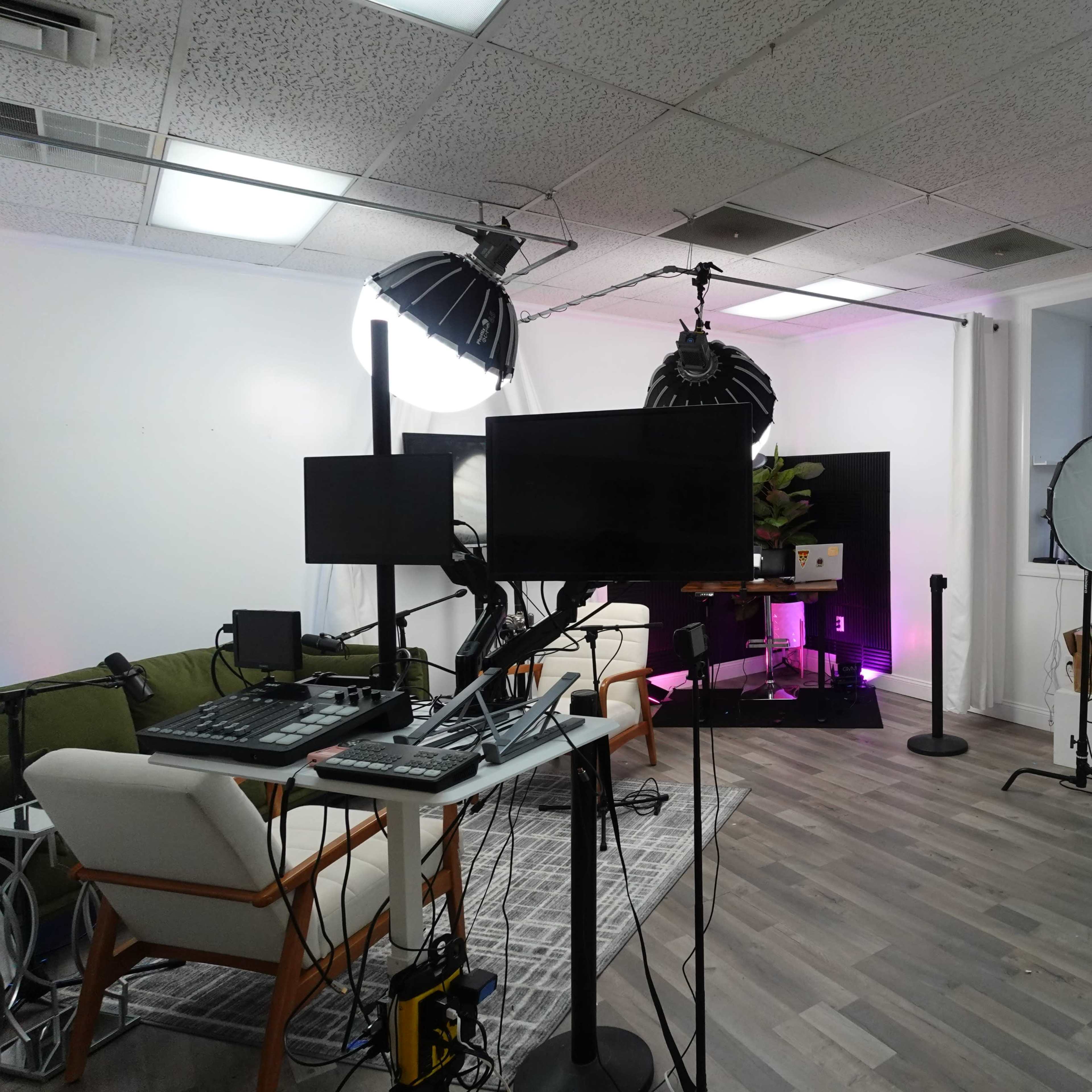 A modern studio space with green plants, multiple screens, lighting equipment, and seating arranged for filming or photography.