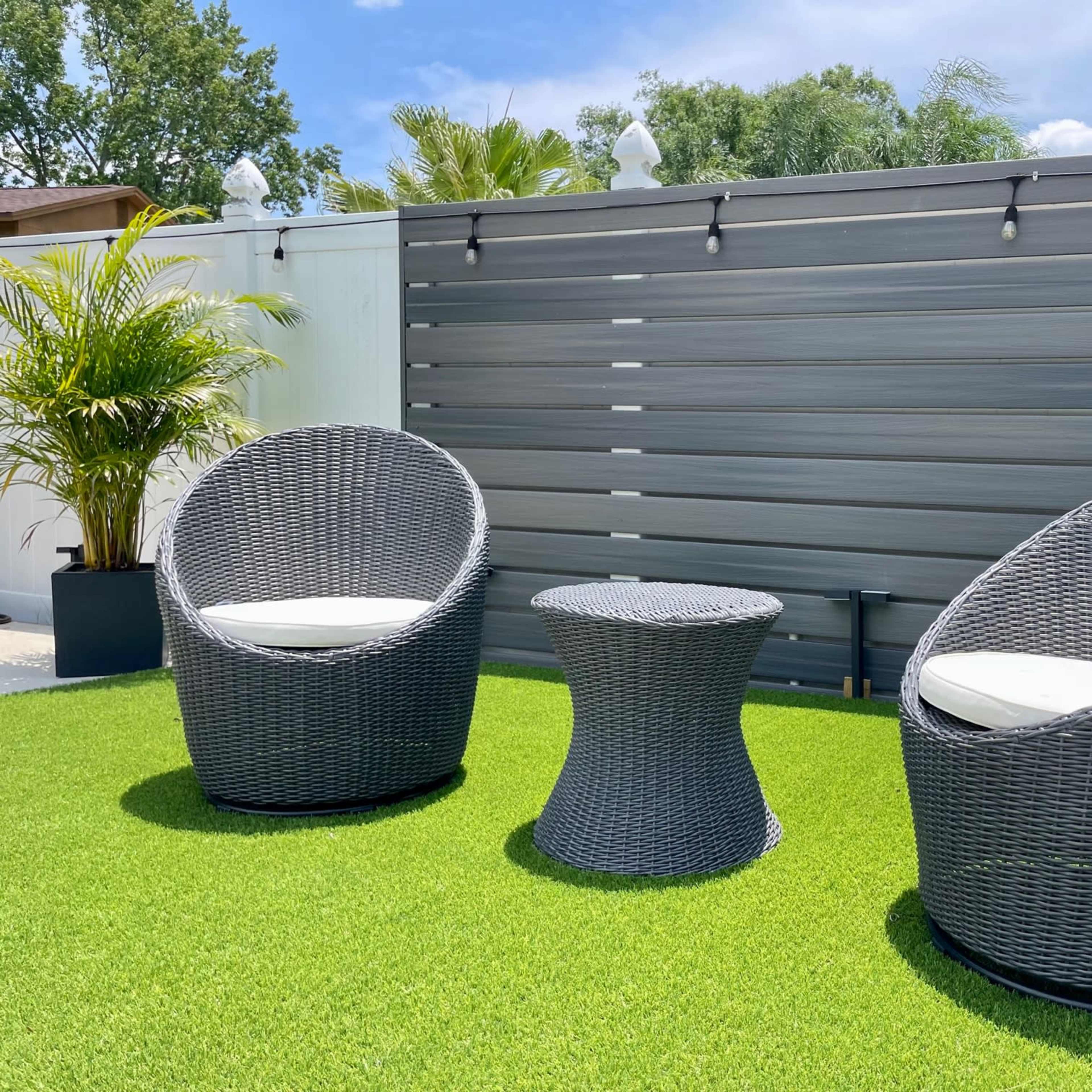 Two round wicker chairs with white cushions and a matching table are arranged on a green grassy area in a backyard setting.