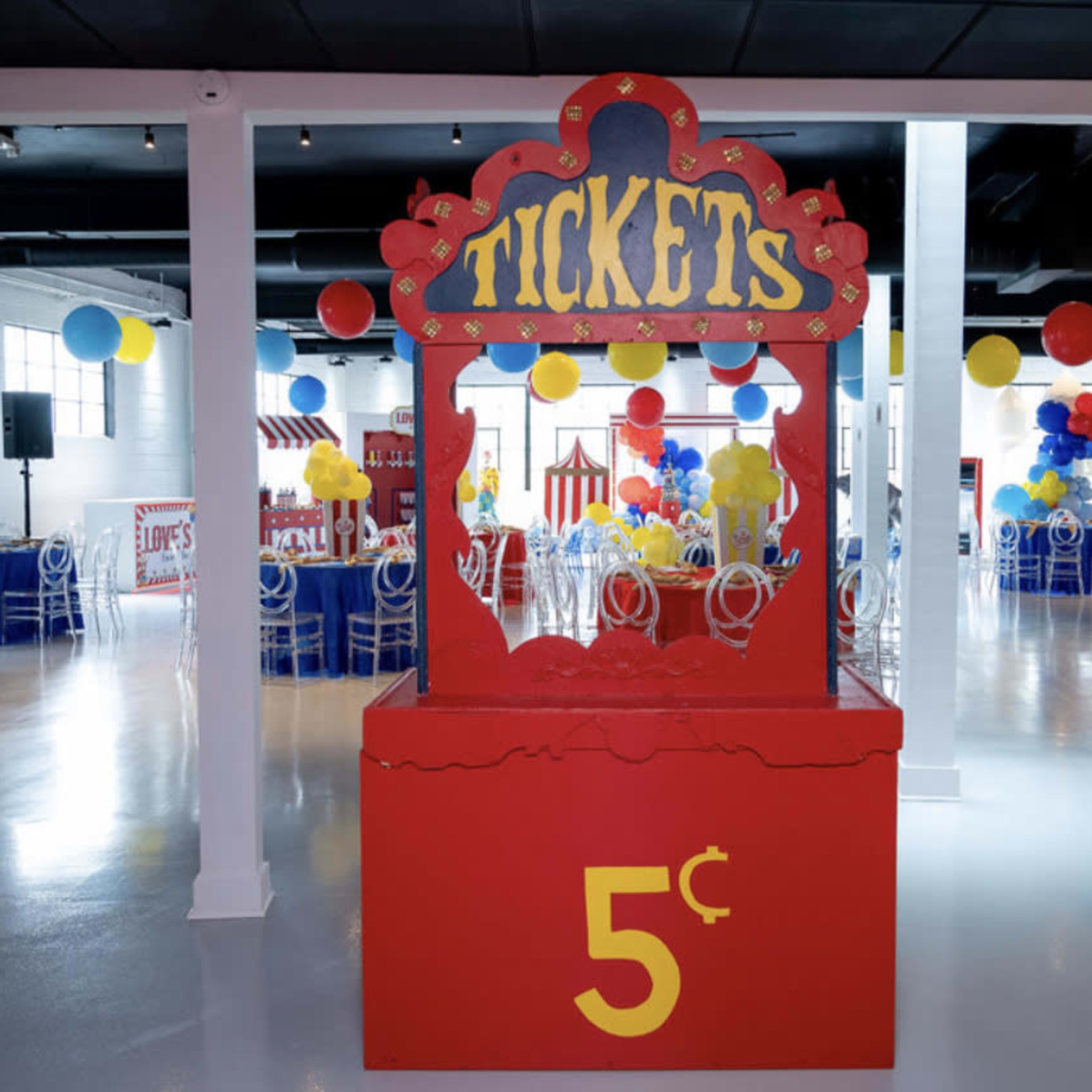 A colorful ticket booth marked with "TICKETS" and a price of 5 cents stands in an event space decorated with balloons and tables.
