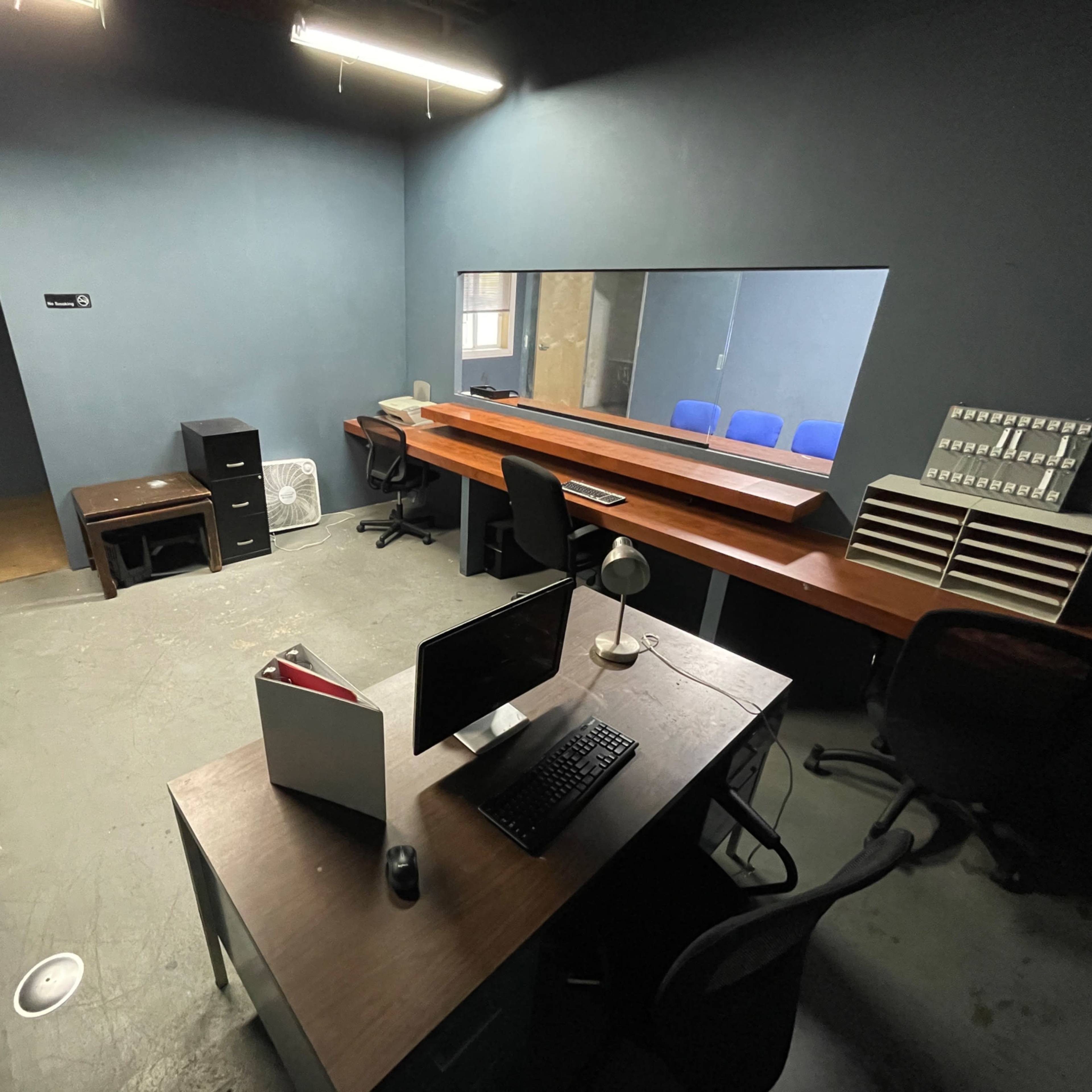 A sparsely furnished office with a desk, computer, filing cabinets, and a long table along the wall, all within a dimly lit space.