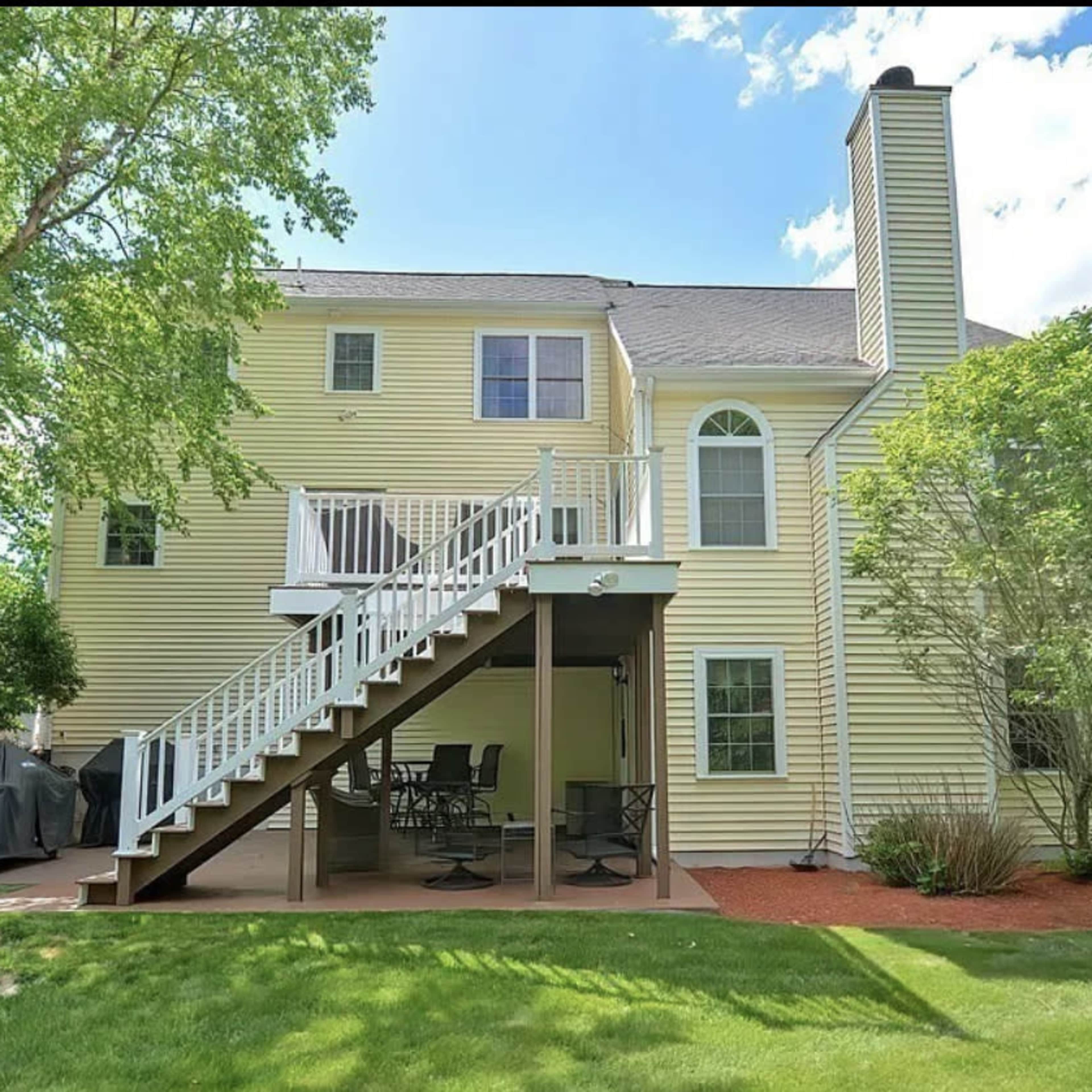 The back exterior of a yellow two-story house with a deck and patio area, surrounded by lush greenery.