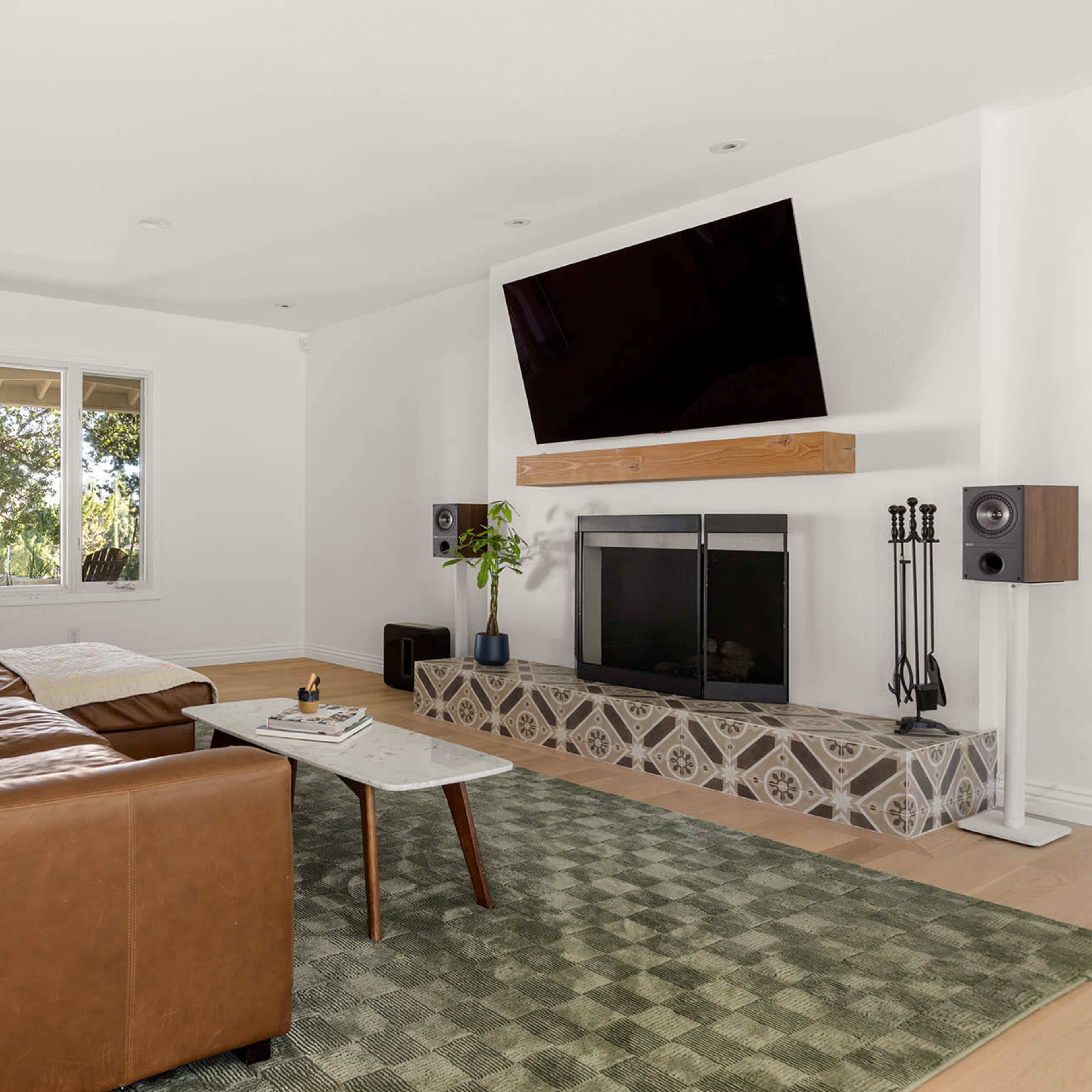 The living room features a brown leather sofa, a wall-mounted TV above a fireplace, and a patterned tile surround.