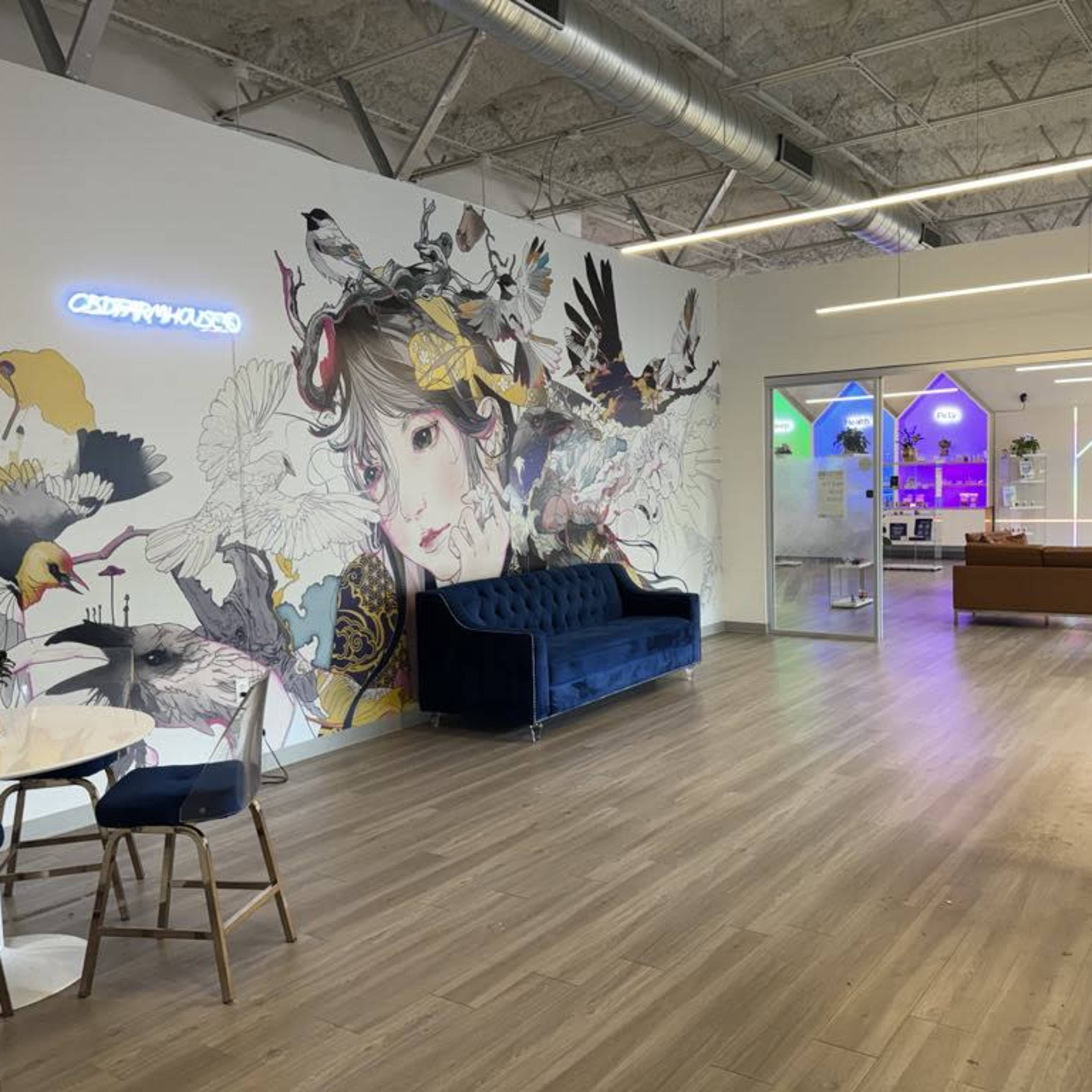 A modern office space featuring a mural of a girl surrounded by birds, with two blue sofas, a round table, and glass walls leading to another area illuminated in colorful lights.