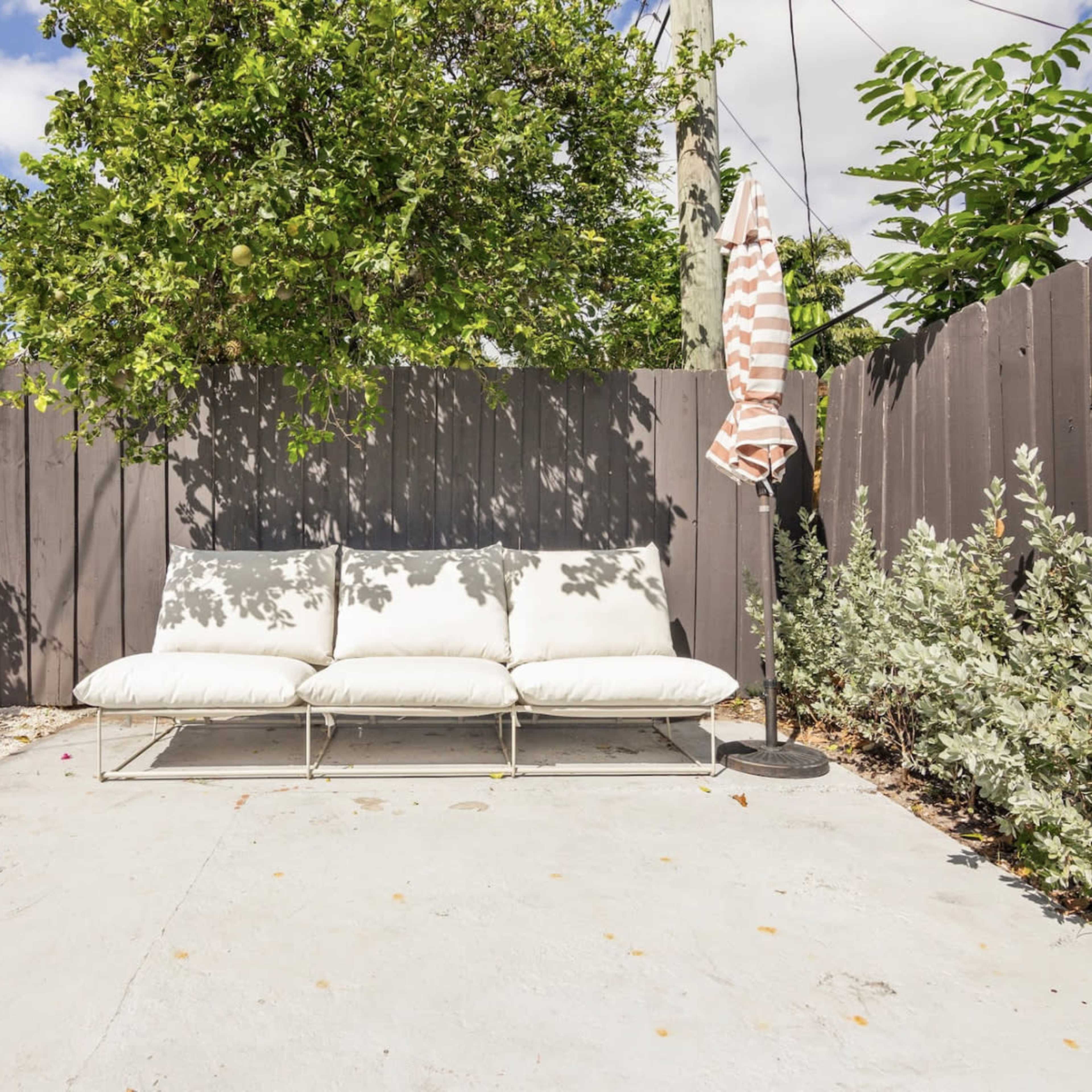 A white outdoor sofa is placed in a patio area surrounded by greenery and a wooden fence, with an umbrella nearby.