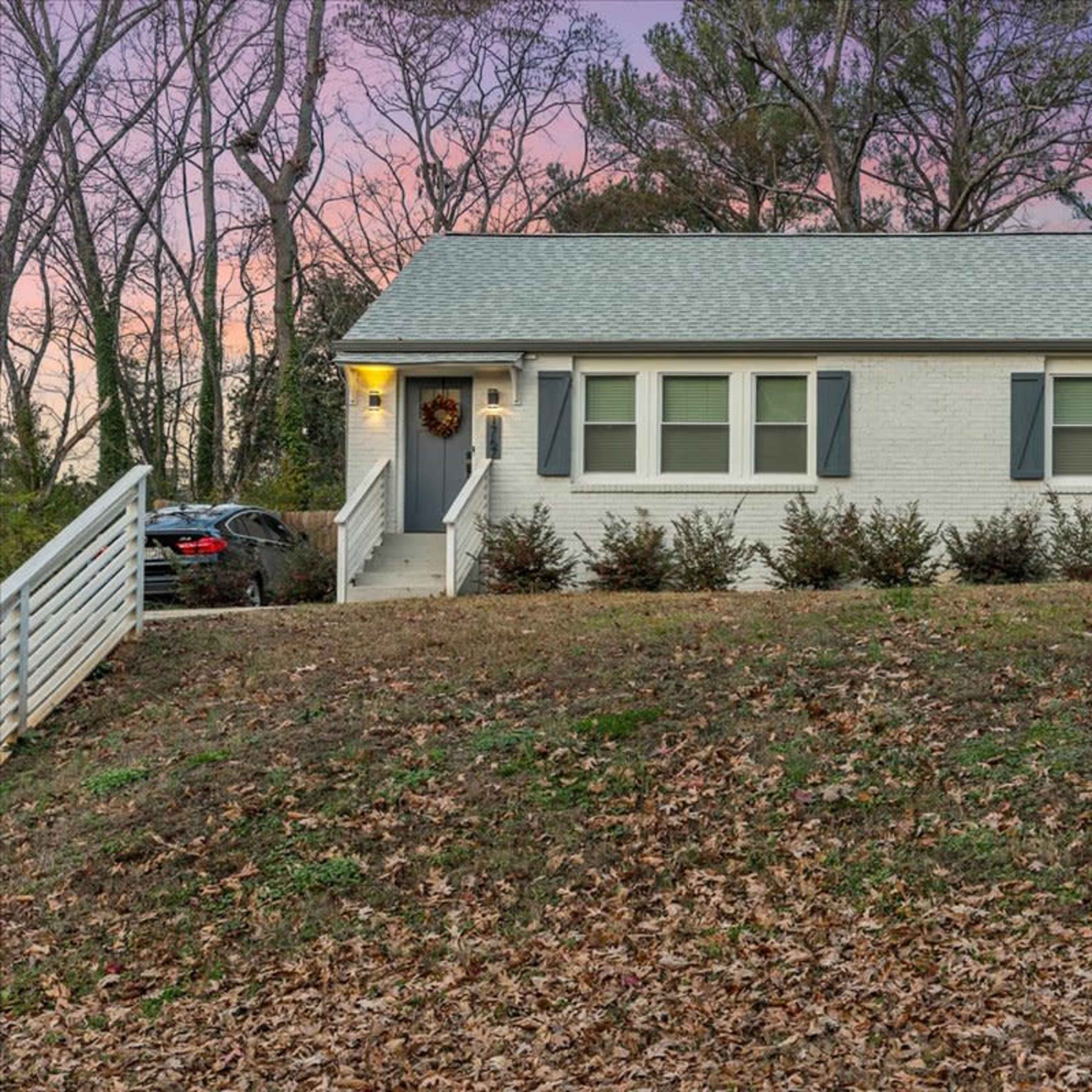 A single-story white brick house sits on a sloped grassy lawn surrounded by trees, with a porch and a visible driveway.