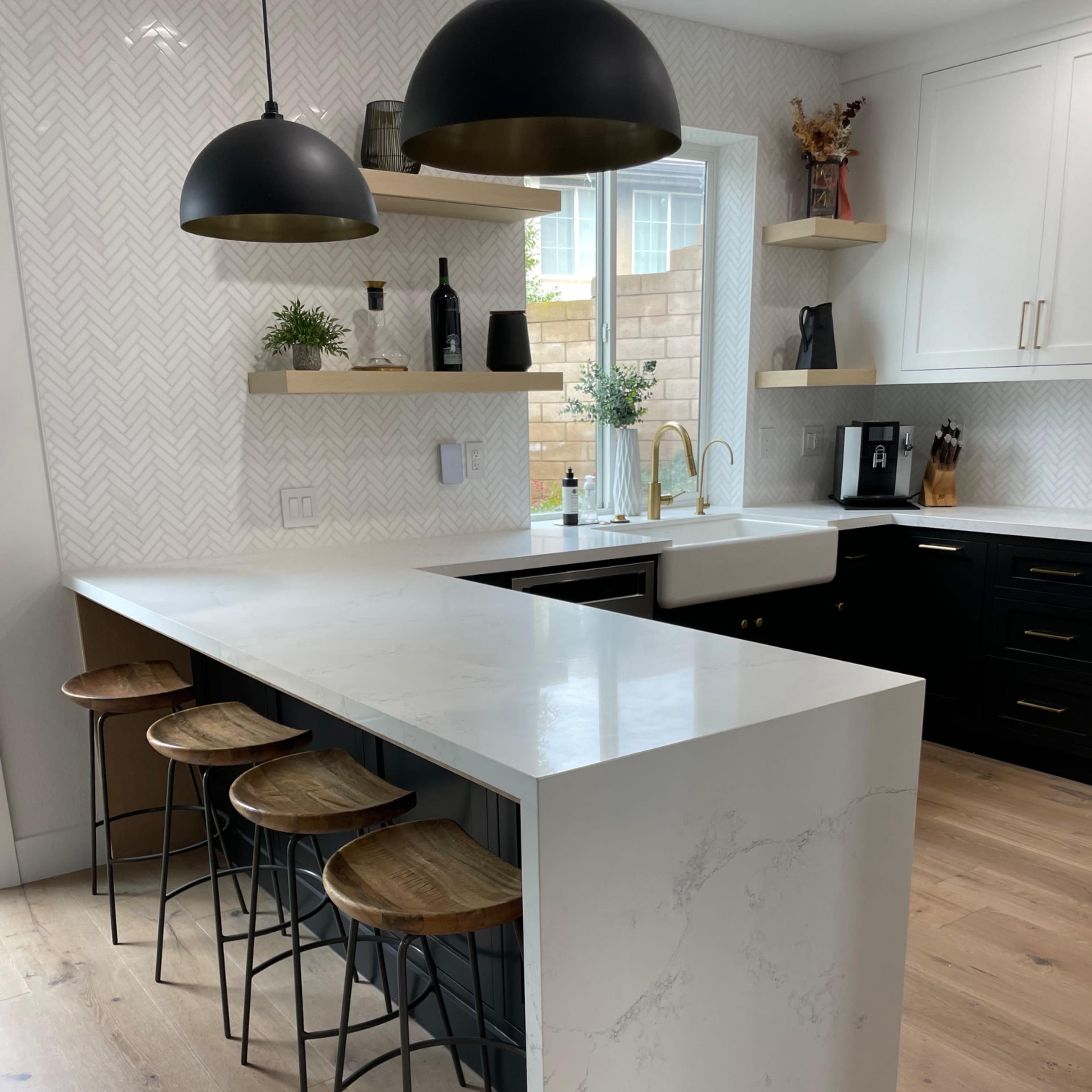 A modern kitchen with a white marble island, three wooden bar stools, black cabinets, and pendant lighting.